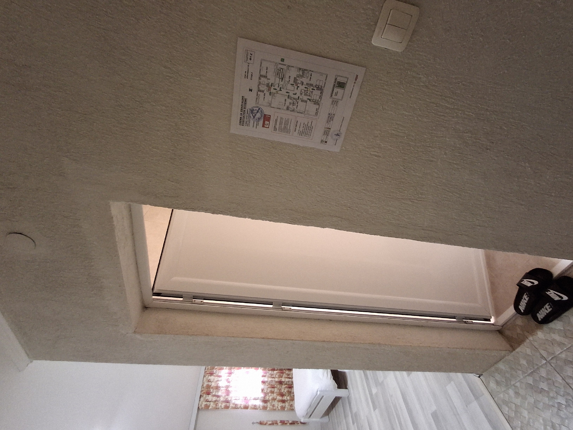 The image depicts a room in Krumovgrad, Bulgaria. The view is looking upwards towards the ceiling, which is textured and off-white in color. A light switch is visible near the top of the frame, along with a printed evacuation plan attached to the ceiling. A rectangular opening in the ceiling leads to another room. Through this opening, a white bed and a window with patterned curtains can be seen. A pair of black Nike-branded slides is on the floor near the opening. The tiles on the floor are light gray.