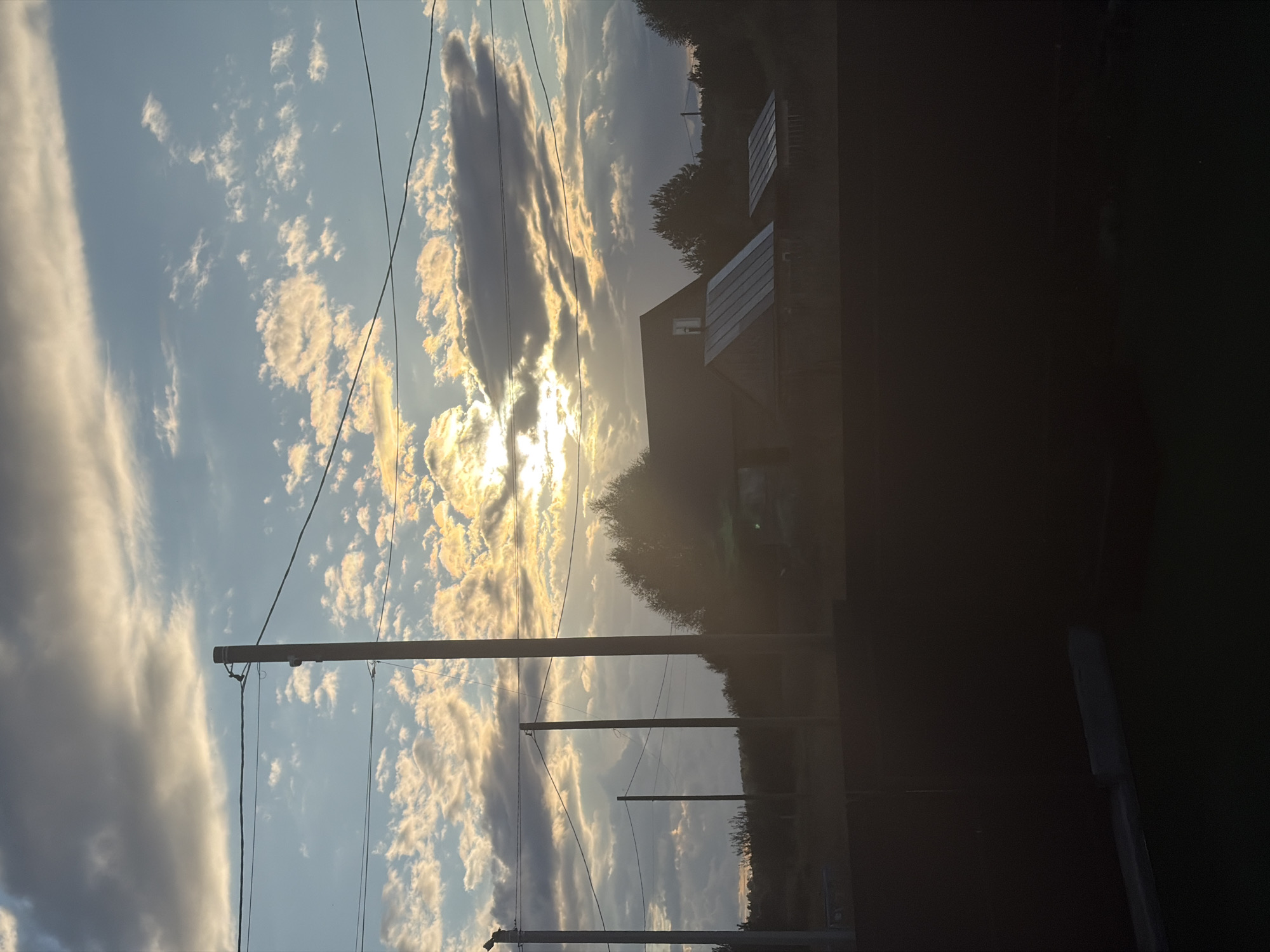 This is an outdoor, eye-level, medium shot of a rural landscape during sunset. The sky is filled with clouds, some dark and some illuminated by the setting sun, which is partially obscured by clouds. In the foreground, there is a dark, silhouetted area that appears to be a fence or wall. Behind this, there are several utility poles with wires stretching across the frame. Further in the background, there are two houses with dark roofs, surrounded by trees. The overall atmosphere is serene, with the warm glow of the sunset casting a peaceful ambiance over the scene. The image does not depict any people or specific identifiable landmarks that would indicate a precise location within Karsha, Russia, other than it being a rural setting.