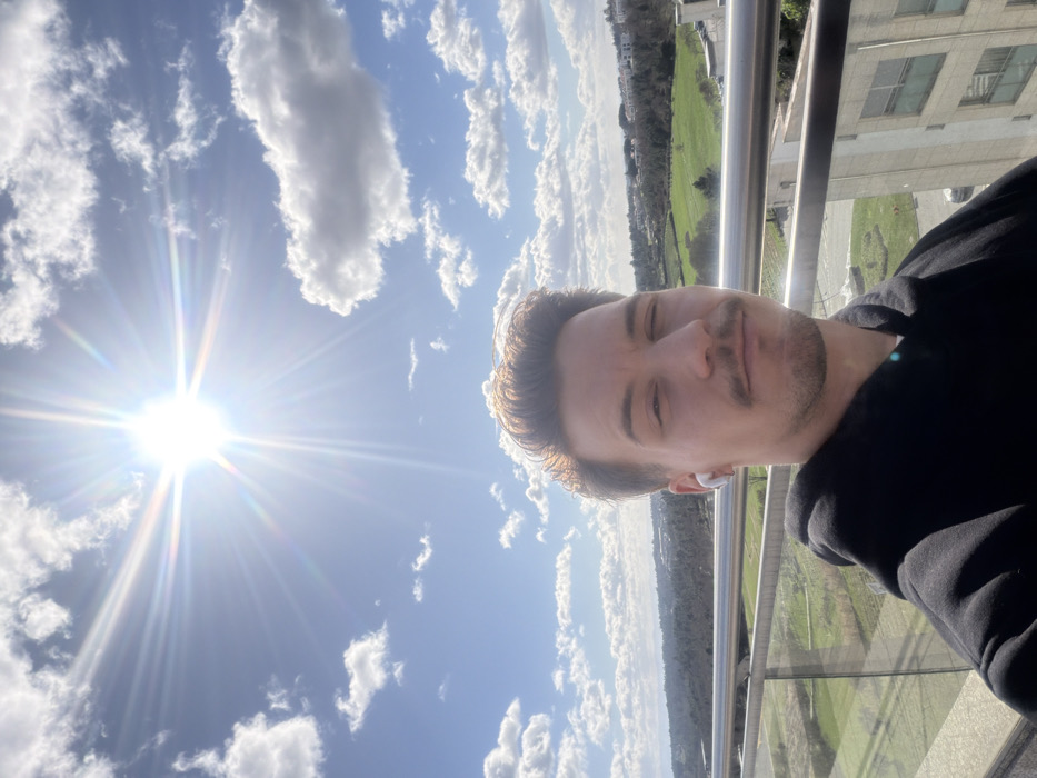 An outdoor portrait, likely a selfie, is taken from a high vantage point, characteristic of a balcony or terrace, in Viseu, Portugal. The primary subject is a male in his 20s or 30s, positioned in the lower half of the frame, looking directly at the camera. He has light brown hair, a mustache, and a short beard. He wears a black hooded sweatshirt and a single white wireless earbud in his left ear. His left eye is partially closed, possibly squinting from the bright overhead sun, which is intensely visible in the upper left quadrant of the image, producing a prominent starburst lens flare against a clear blue sky with scattered white cumulus clouds. Behind the subject, a modern glass and metal railing extends horizontally. The background reveals a panoramic view of Viseu, encompassing rolling green hills, dense tree cover, cultivated land, and several multi-story buildings with light-colored facades.