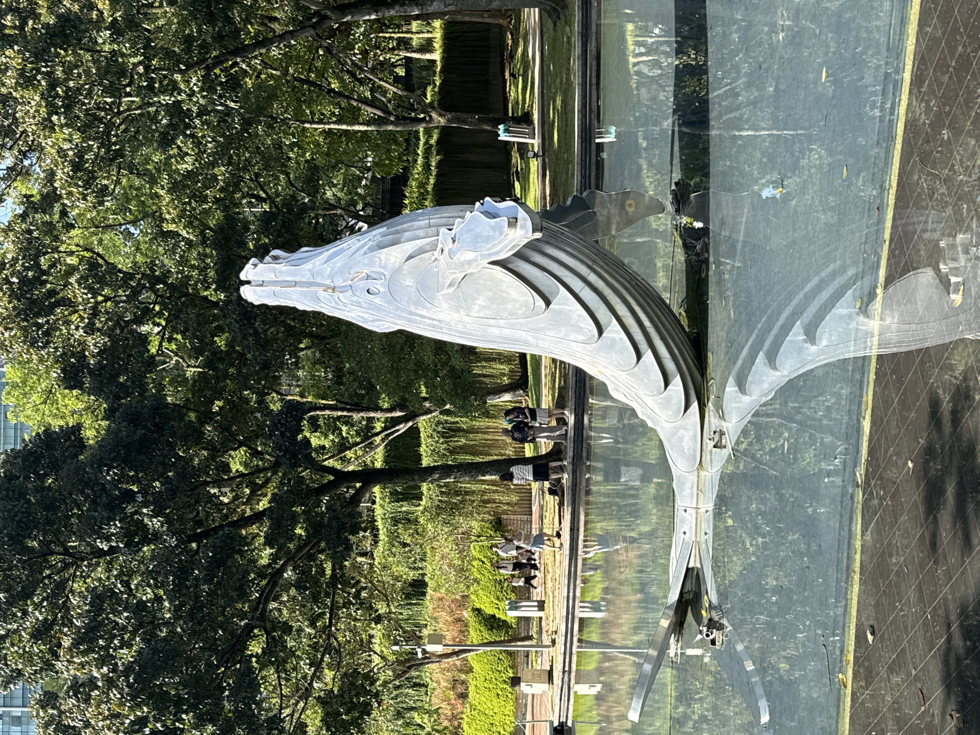 The image shows a silver, stylized metal sculpture of a horse's head and neck with a flowing mane. The sculpture is positioned in front of a large window, creating reflections of an outdoor scene. The outdoor scene includes green foliage, trees, and a sidewalk with people walking. The window has a dark, tiled frame or border. The scene is Kuala Lumpur, Malaysia.