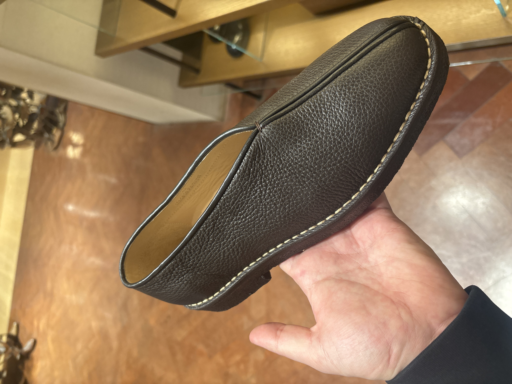 A person is holding a dark brown leather slipper with light stitching around the sole. The slipper has a simple design with a rounded toe and a slightly raised seam along the top. The interior lining is a light tan color. The person is wearing a dark colored long sleeve shirt. The background includes a blurred view of a room with a display shelf and a reddish-brown tiled floor. The location is not identifiable as London, United Kingdom from the visual information.
