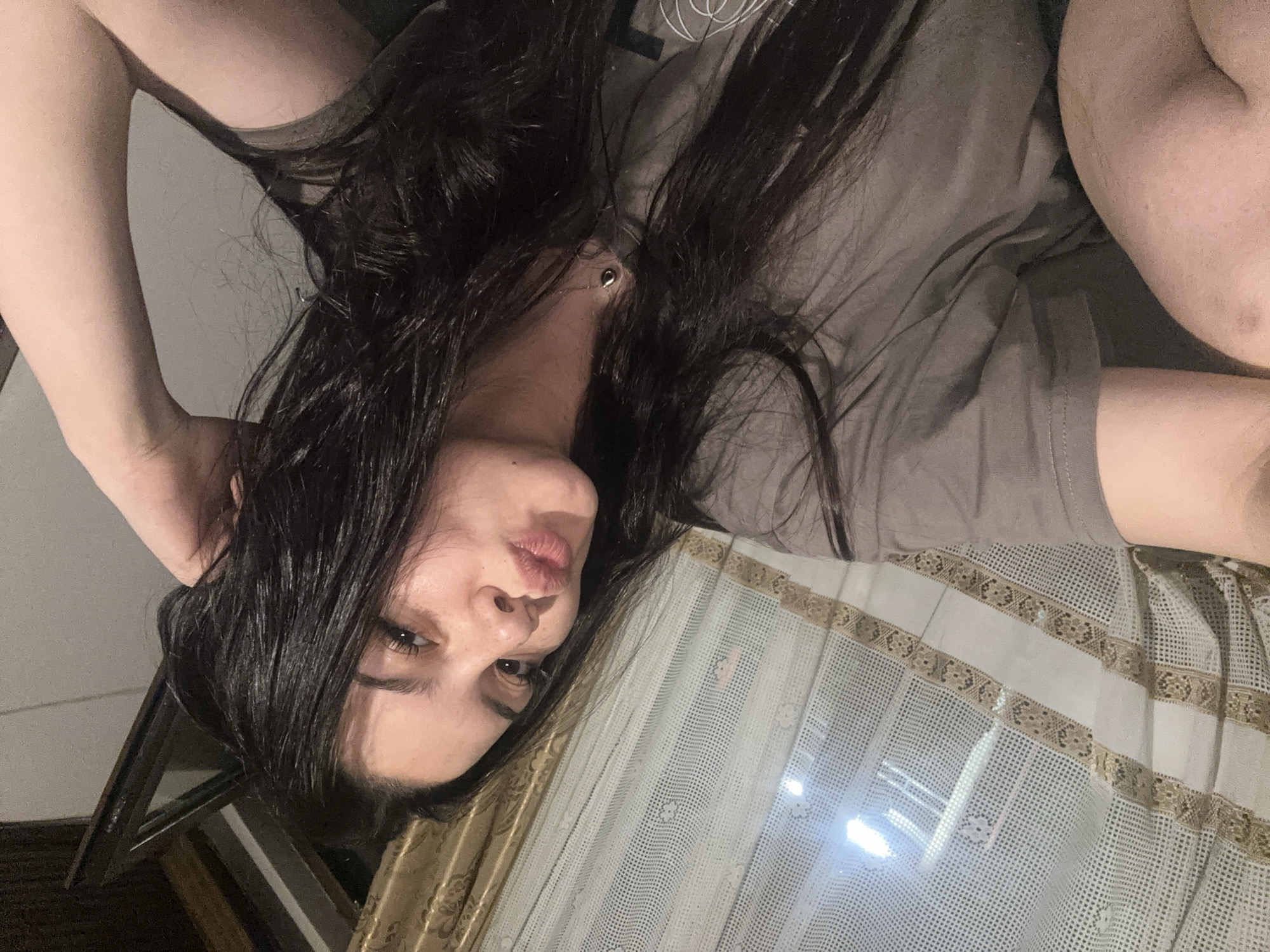An indoor portrait shows a person with long, dark hair and fair skin, wearing a plain grey t-shirt. The individual is positioned as if lying down, with their head tilted and one eye looking directly at the camera. Their lips are slightly pursed in a soft expression. One arm is bent, with the hand near the back of their head, partially obscured by hair. A small, gold-colored pendant or earring is visible near their neck, nestled within their dark hair.
The background on the left is dark and indistinct, likely a wall or furniture. On the right side of the frame, a significant portion is occupied by a textured fabric, appearing to be a curtain or decorative textile. This fabric features a white, open-weave or lace-like pattern, horizontally striped with ornate gold-brown bands. Below this fabric, a dark, possibly wooden or metallic frame of a picture or mirror is visible. The lighting is artificial, with bright reflections on the patterned fabric suggesting an internal light source. The overall scene conveys a relaxed atmosphere within a domestic setting, possibly a bedroom or living area in Chubra, Bulgaria. No text is visible in the image.