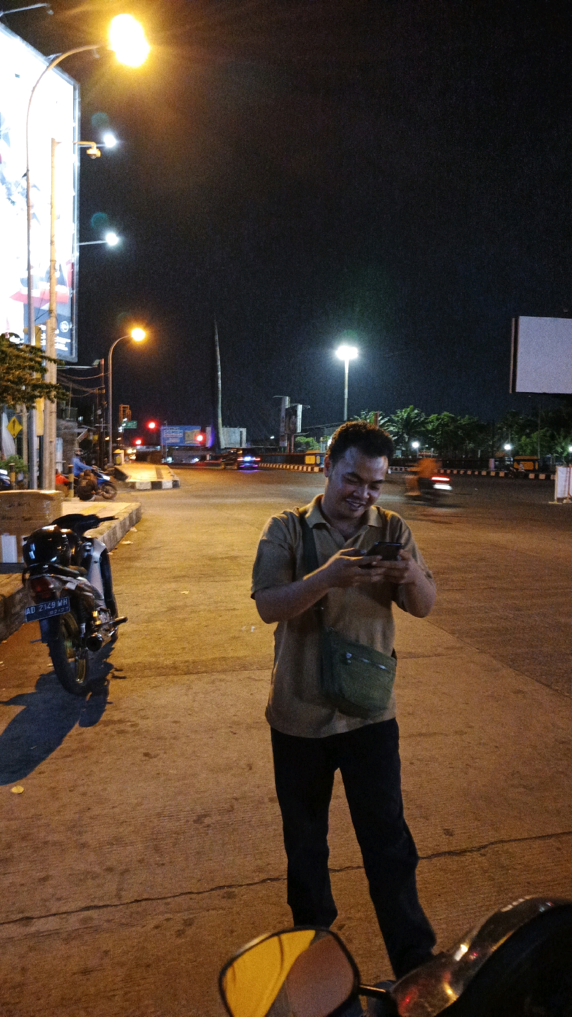 A man, of Indonesian descent with dark hair and a light complexion, is standing on a street in Surakarta, Indonesia at night. He is wearing a tan collared shirt, dark pants, and a green satchel bag slung across his chest. The man is looking down and smiling while holding and interacting with a smartphone.
He is positioned on a concrete sidewalk adjacent to a road with streetlights illuminating the scene. Several motorcycles are visible, including one parked to his left and another in motion further down the road. Traffic lights and other vehicles are present in the background, indicating an active urban environment. The overall atmosphere is illuminated by artificial lights, creating a warm and somewhat grainy image.