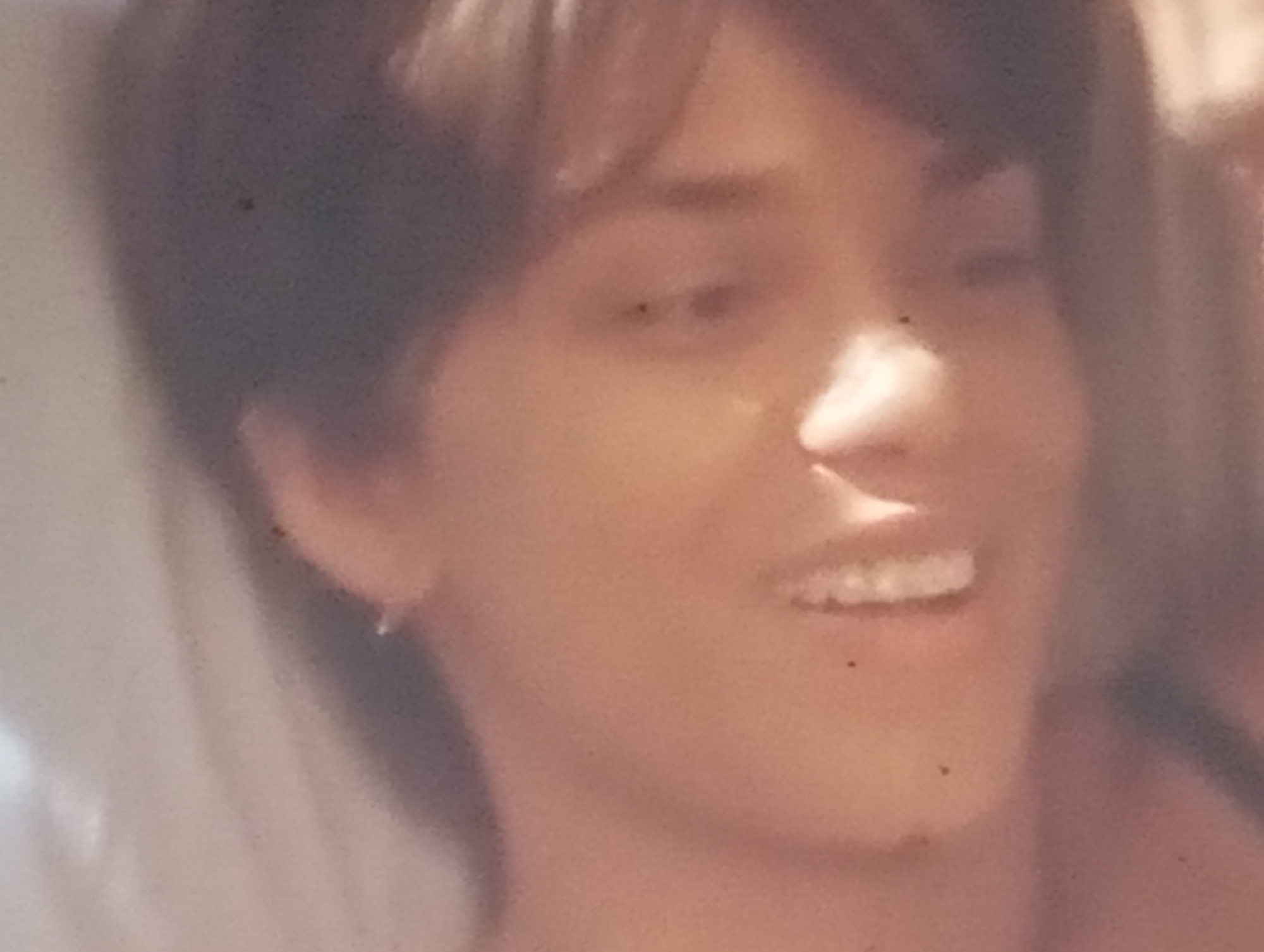The media file is a close-up, softly focused portrait of a young woman. She has short, dark hair and appears to be smiling. Her eyes are open, and her mouth is slightly parted, revealing her teeth. She is wearing a small hoop earring in her left ear. The lighting suggests an indoor setting, possibly during the day, as there is a bright light source illuminating her face from the side. The overall tone of the image is warm and intimate, conveying a sense of happiness or amusement. There is no visible text or specific location cues beyond the provided context of Sofiivska Borshchahivka, Ukraine.