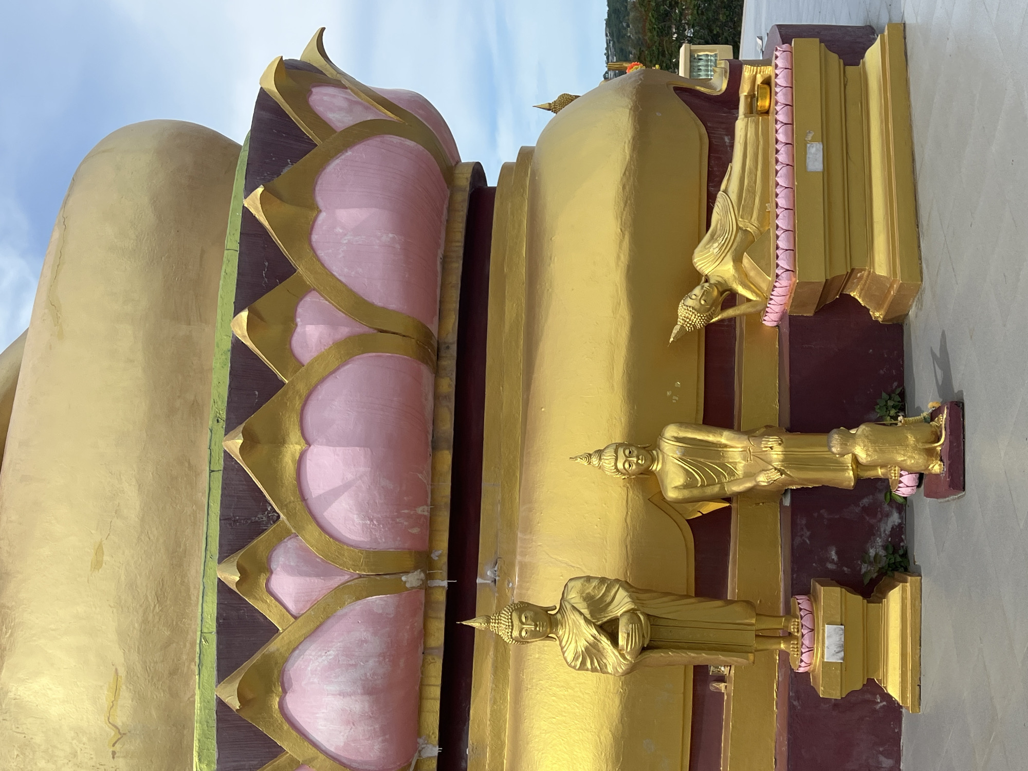 In Baan Plai Laem, Thailand, this image captures the base of a large, golden stupa adorned with pink lotus petals outlined in gold. In front of the stupa, three golden Buddha statues are displayed. A standing Buddha statue, arms crossed in meditation, is on the left. To its right, a smaller standing Buddha statue is positioned with hands clasped at its chest, and a golden monkey kneels at its feet. The furthest right statue depicts a reclining Buddha, head supported by its hand, lying on a golden pedestal with pink lotus embellishments. The ground is paved with grey concrete. The sky is partly cloudy.