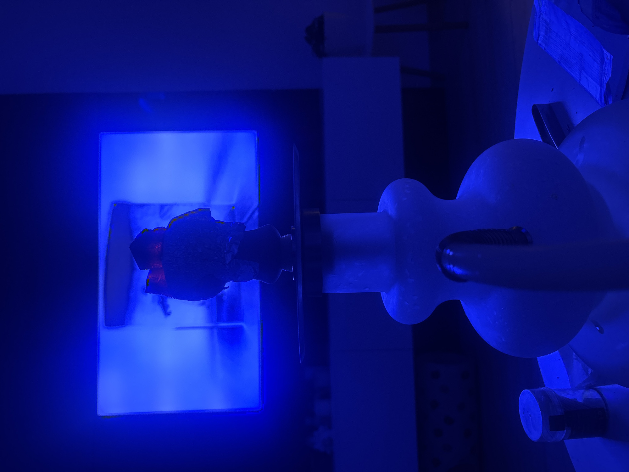 The image depicts a hookah in the foreground, with a television screen displaying a bright blue image in the background. The scene is bathed in blue light, suggesting ambient lighting or a specific mood setting. The hookah is a frosted glass base with a dark bowl on top, covered in what appears to be aluminum foil and charcoal. The television screen is large and emits a strong blue glow. The overall environment appears to be indoors, possibly a living room or entertainment space, with furniture visible in the background, partially obscured by the dim lighting. There are no people visible in the image. The composition focuses on the hookah and the illuminated television screen.