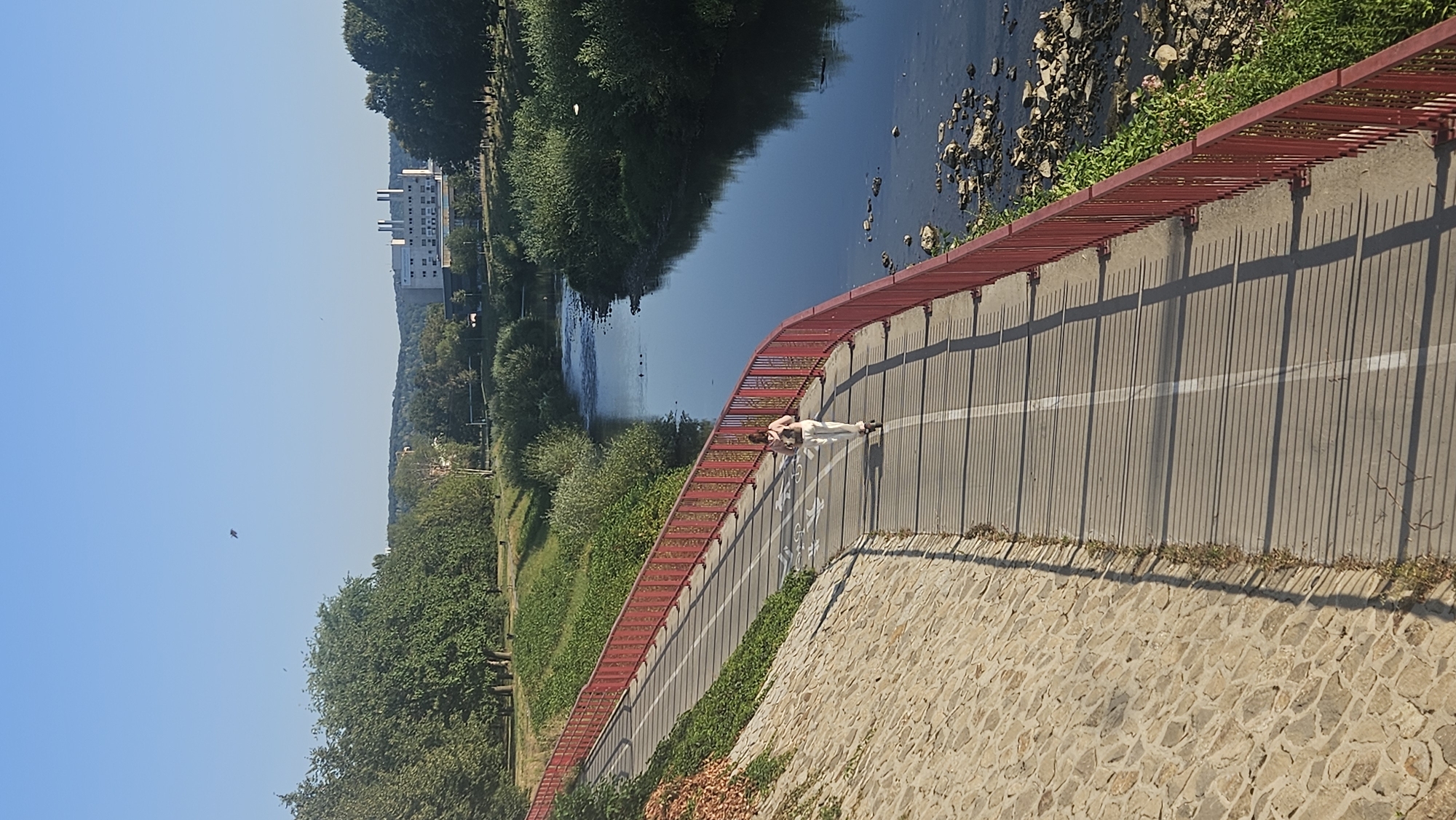 A young person rides a scooter on a paved path next to a red metal railing that separates the path from a river. The person is wearing a light-colored top and light-colored pants. The path has white lane markings, and bicycle symbols are painted on it. The river is dark blue and has rocks along its bank. Green trees and vegetation are visible on the opposite bank and in the background. A large, white building with multiple chimneys stands in the distance, situated on a hill among trees. The sky is clear and blue, with a bird flying high up. The scene appears to be a park or recreational area within the city of Prešov, Slovakia, likely near the Torysa River.