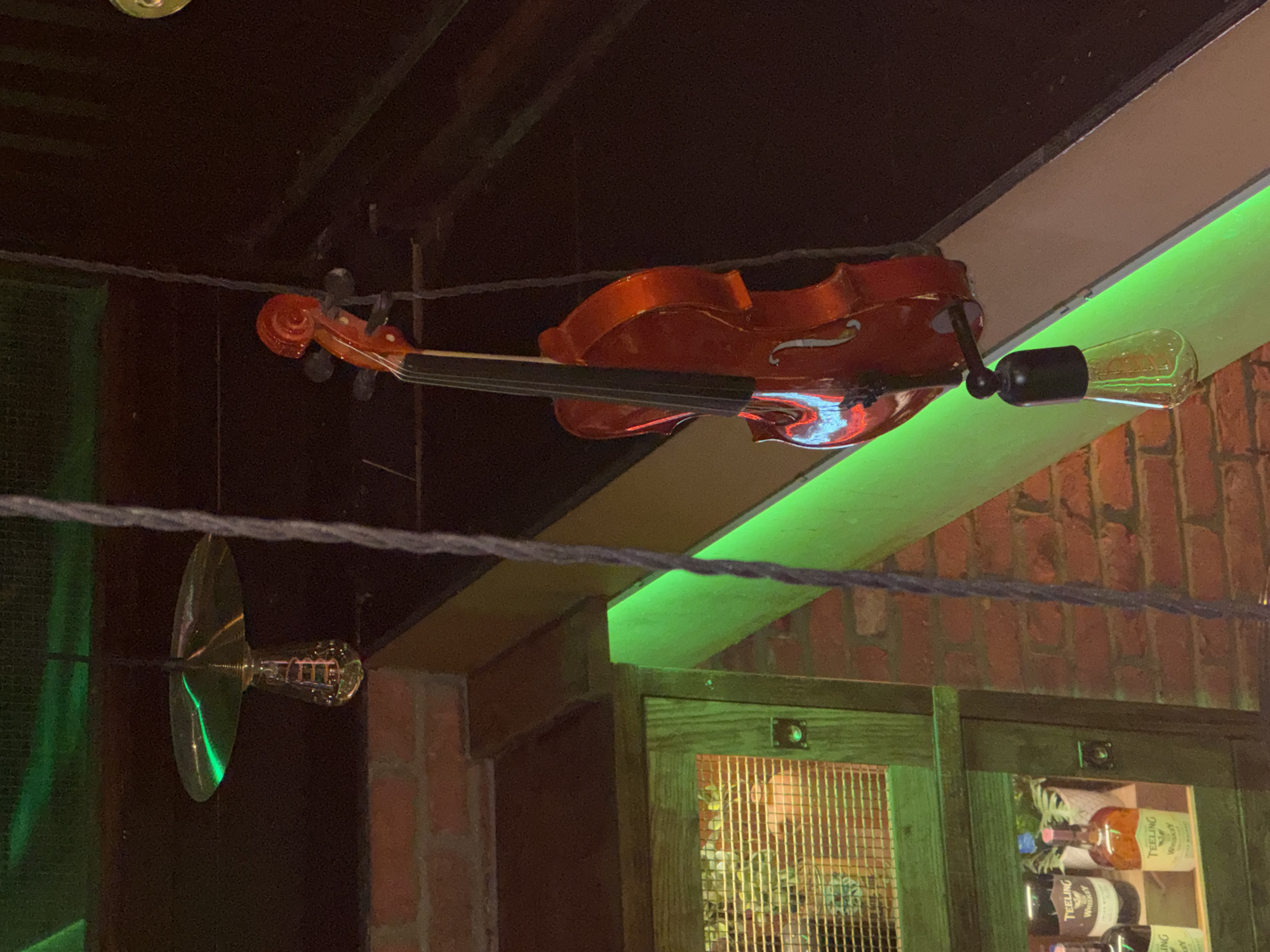 The image presents a dimly lit interior scene, likely a bar or pub in the evening, characterized by its decorative elements and atmospheric lighting. A vibrant orange-red violin is suspended horizontally from the ceiling, serving as a prominent decorative feature in the upper part of the frame. An Edison-style lightbulb is attached to the right side of the violin, emitting a warm light, and another similar bulb is visible to the left, reflecting the ambient green illumination.
Dark cables or wires crisscross the upper and middle sections, from which the violin and lightbulbs are suspended. A striking green LED light strip casts a vivid glow along a wooden beam or shelf beneath the violin. Below this, an exposed brick wall forms a rustic backdrop.
In the lower right, a dark wooden cabinet or shelving unit with glass or mesh doors is partially visible. Inside, several bottles of spirits are stored, with the brand "TEELING WHISKEY" clearly legible on at least two bottles. The scene is devoid of people, focusing entirely on the detailed decor and lighting that contribute to a cozy and inviting ambiance, typical of a commercial establishment in London. No specific activity or event is taking place.