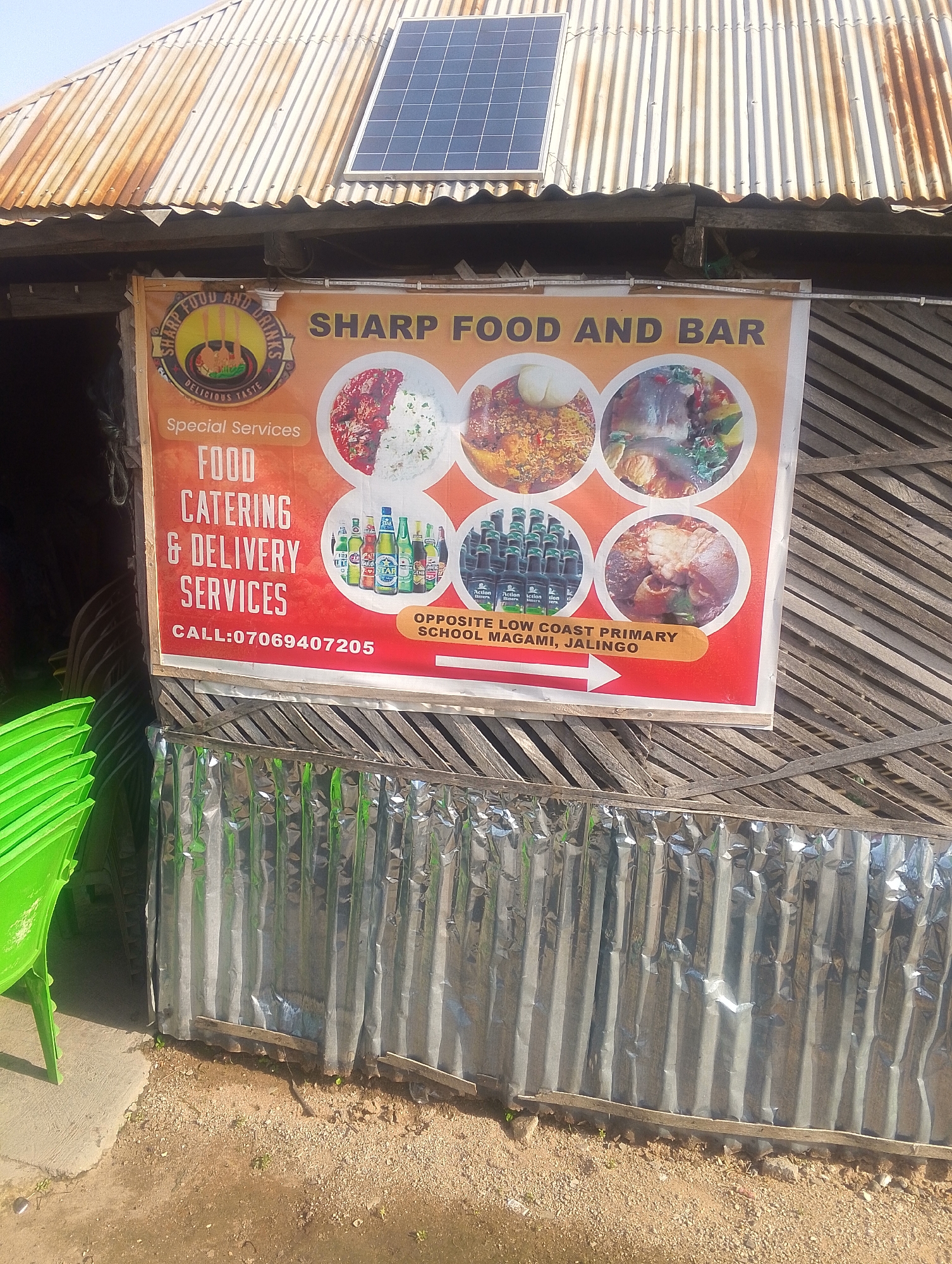 An outdoor, medium shot captures a sign for "SHARP FOOD AND BAR" positioned above a corrugated metal facade. The sign, predominantly orange and red, features images of food and drinks, and lists "FOOD CATERING & DELIVERY SERVICES" and a phone number: 07069407205. Below the phone number, an arrow points to text indicating the location: "OPPOSITE LOW COAST PRIMARY SCHOOL MAGAMI, JALINGO". A solar panel is visible on the corrugated metal roof above the sign. To the left of the sign, a stack of bright green plastic chairs is partially visible. The environment appears to be an outdoor establishment with a rough, unpaved ground surface. The overall scene suggests a small business operating in a local market or roadside setting in Jalingo, Nigeria.