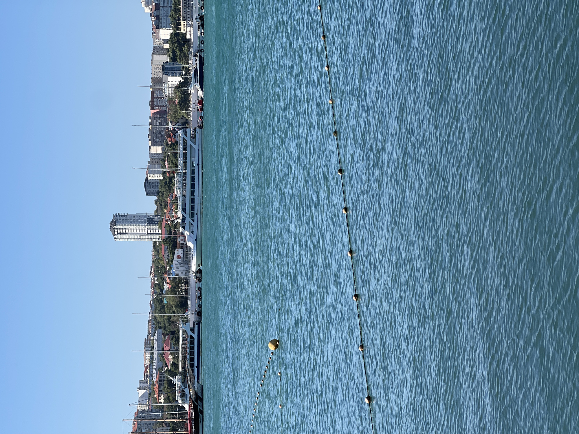A wide-angle, eye-level, outdoor, daytime, vertical shot captures a serene view of the ocean and a coastal city skyline. The turquoise water fills the foreground, gently rippling under a clear blue sky. Two parallel lines of small, light-colored buoys stretch across the water, connected by thin ropes. A larger, yellow buoy is positioned between these lines in the middle-left of the frame.
In the background, a collection of buildings forming a city skyline are nestled against a backdrop of lush green trees. The buildings vary in height and architectural style, with a prominent tall, white, modern high-rise standing out against the more traditional structures. Several boats, including yachts and larger passenger vessels, are docked along the waterfront, their masts and superstructures visible. The overall scene evokes a sense of tranquility and urban coastal life.
