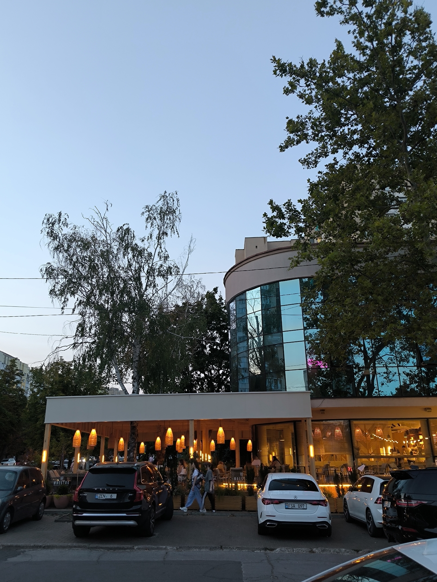 In Chișinău, Moldova, an outdoor seating area of a restaurant, lit by hanging, cone-shaped lamps, is visible. Several cars are parked in front of the restaurant, including a black Volvo XC90 and a white Mercedes-Benz. A modern building with a curved, glass facade stands behind the restaurant. Trees with green foliage are scattered throughout the scene, with the sky a clear, pale blue.