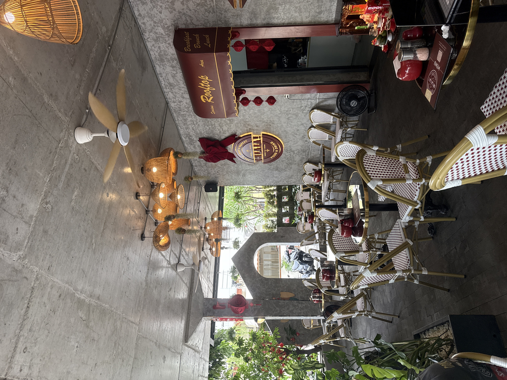 This appears to be an indoor/outdoor restaurant scene. The restaurant, called "Lalati," has an outdoor "Rooftop" area for Breakfast, Brunch, and Lunch. The interior design features rattan lighting fixtures and a modern concrete wall. The restaurant has numerous tables and chairs, and a menu is visible on one table. The scene is not crowded, but some people are present in the background, near a gateway that offers a view of greenery, possibly a garden or patio. The exact location is unknown, but given the name, it could be in a city.
