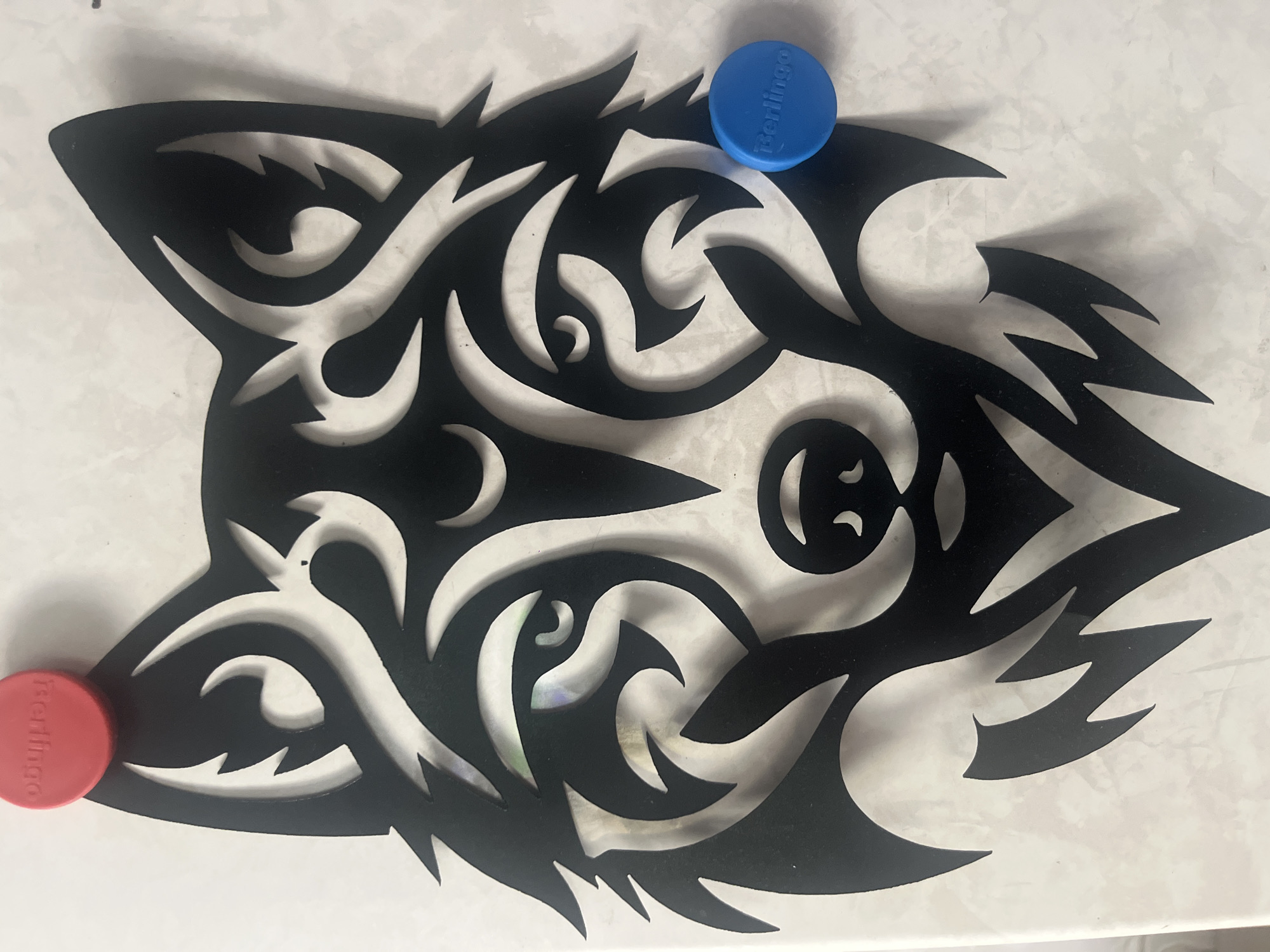 The image shows a black, stylized wolf head cutout attached to a white, textured surface, likely a wall or whiteboard. There are two small, round magnets on the surface: a red one near the top left of the wolf's head, and a blue one near the right eye. The red magnet has the word "Berlingo" printed on it in white, reversed text. The blue magnet also has the word "Berlingo" printed on it in white text. The wolf design features sharp, angular lines and tribal-like swirls. The surface appears to be indoors and well-lit. There is no discernible location in the city of Tyumen, Russia.