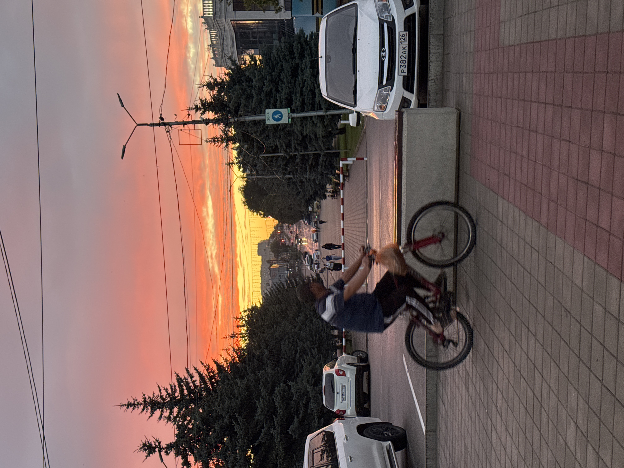 A man rides a bicycle on a paved sidewalk in Mikhaylovsk, Russia, during sunset. The sky is ablaze with vibrant hues of orange, pink, and yellow. Tall evergreen trees line the street, and a white car is parked nearby. In the background, buildings and streetlights are visible, and other people can be seen walking.