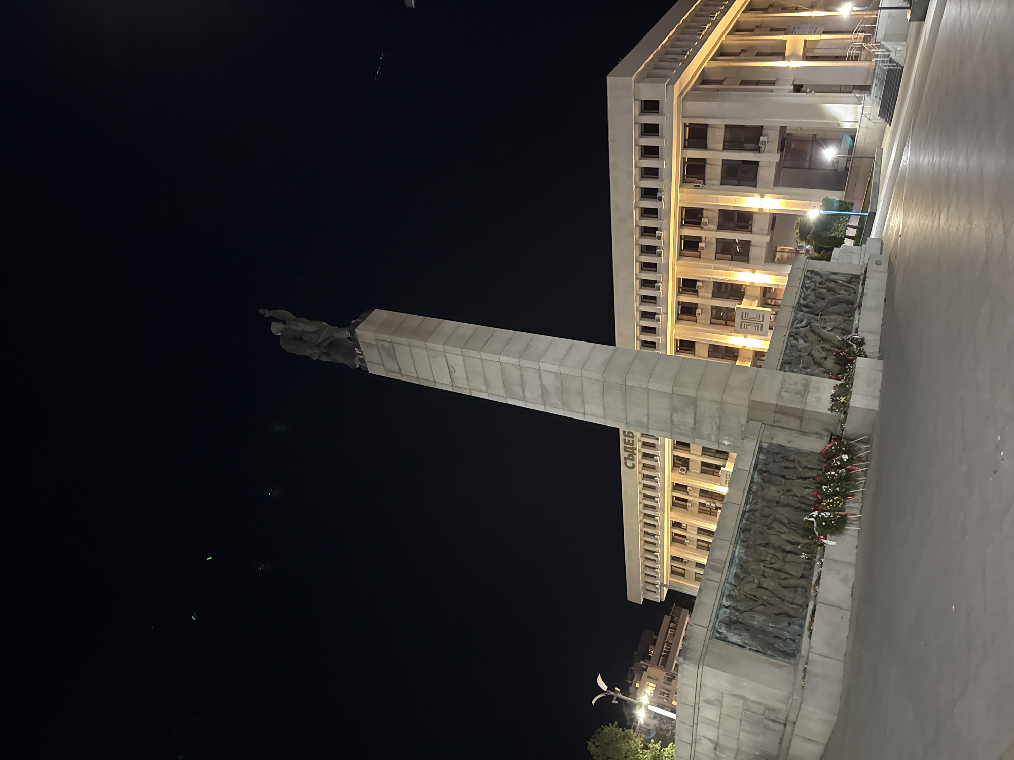 The image depicts a night scene in Burgas, Bulgaria, showcasing the Soviet Army Monument and a portion of the Burgas Municipality building. The monument is a towering concrete structure, topped with sculpted figures of soldiers. The base of the monument features bas-reliefs of figures engaged in various activities, possibly depicting scenes of war or labor. Floral wreaths are placed at the base, suggesting commemorations or memorials. The Burgas Municipality building, a multi-story structure, is visible in the background, with rows of illuminated windows. The sky is dark, indicating nighttime. A street lamp illuminates the area around the monument. The ground appears to be paved, possibly a pedestrian area or square.