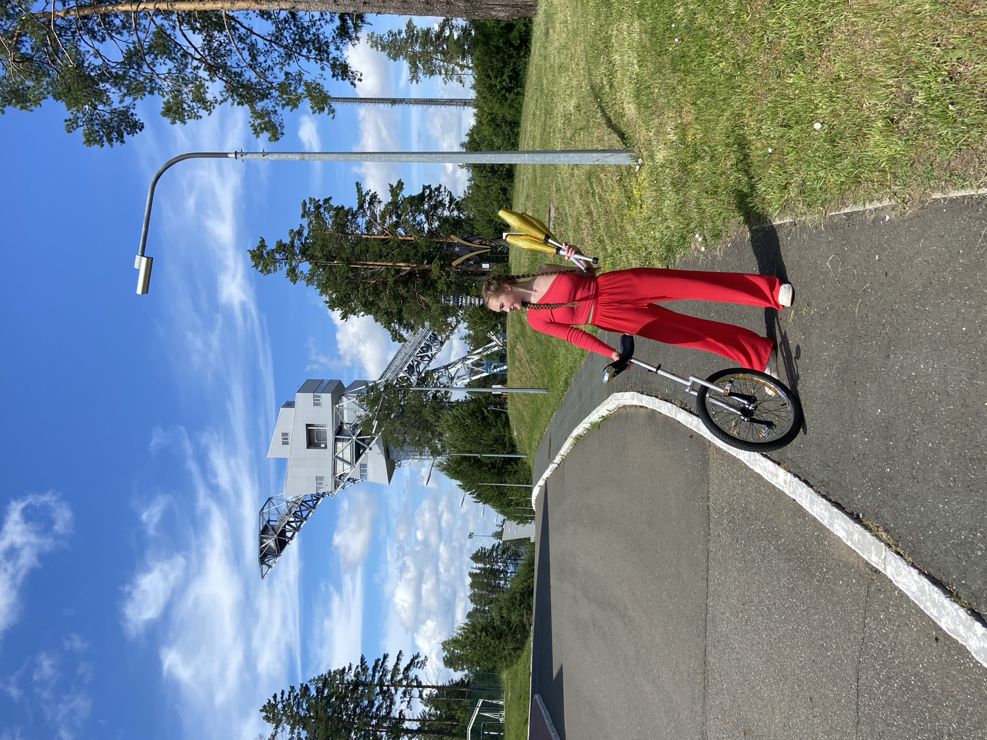 A young Caucasian female stands on a paved walkway next to a road, holding juggling clubs and beside a unicycle. She is wearing a red one-shoulder crop top and loose red pants. The background features a large, modern ski jump structure under a bright blue sky with scattered white clouds. Green trees and grass are visible on either side of the road and walkway. A tall lamppost stands to the right of the woman. The scene appears to be set in a park or recreational area. The location is specified as the city of Prikamsky, Russia, at the Chaykovsky ski jumping complex.