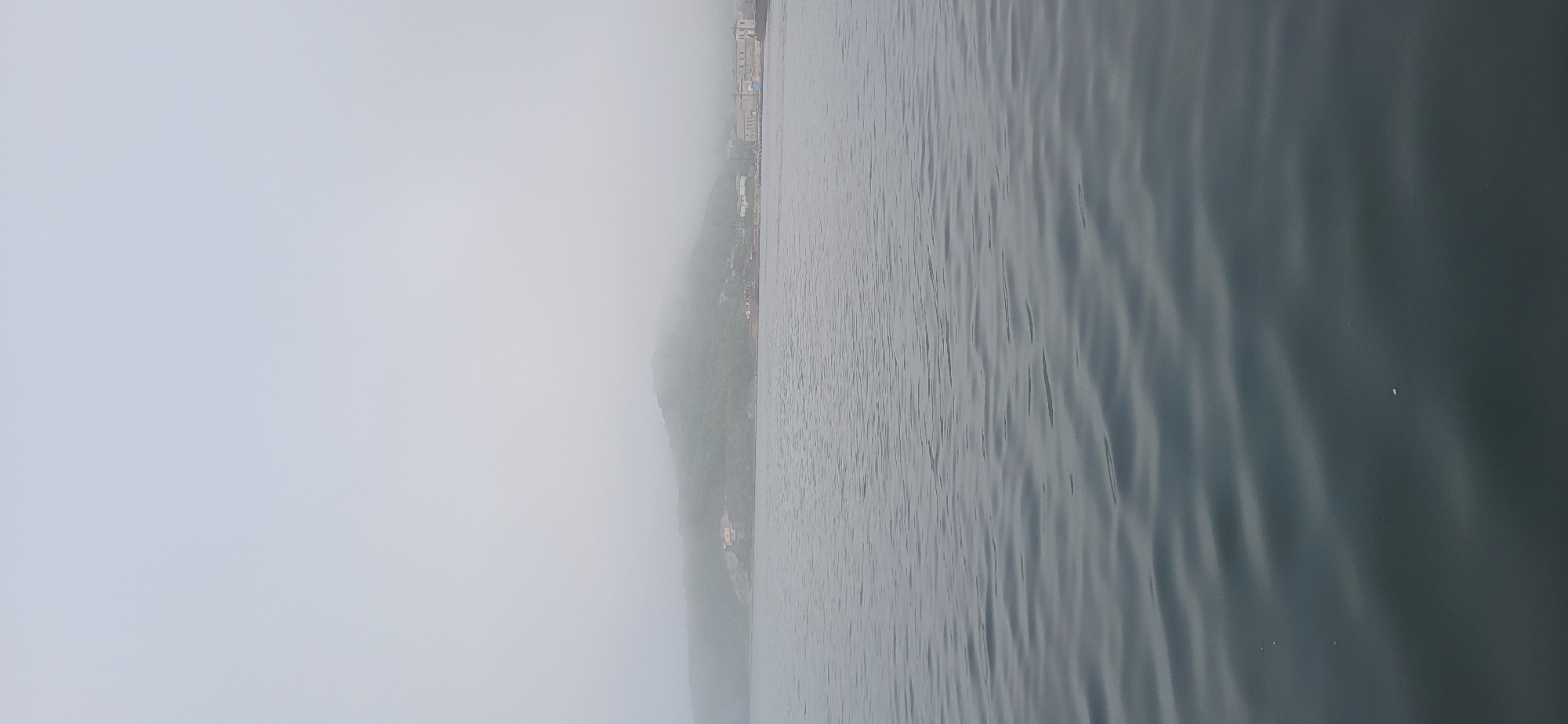 The image captures a serene, overcast day in Slavyanka, Russia, with a tranquil body of water occupying the foreground. The water's surface is gently rippled, reflecting the muted, diffused light of the sky. In the background, a verdant, mist-shrouded hillside rises from the coastline, its contours softened by the fog. Along the base of the hill, a few buildings are visible, suggesting a small settlement or coastal town. The overall atmosphere is one of quietude and natural beauty, characteristic of a foggy day by the sea in this coastal Russian city.
