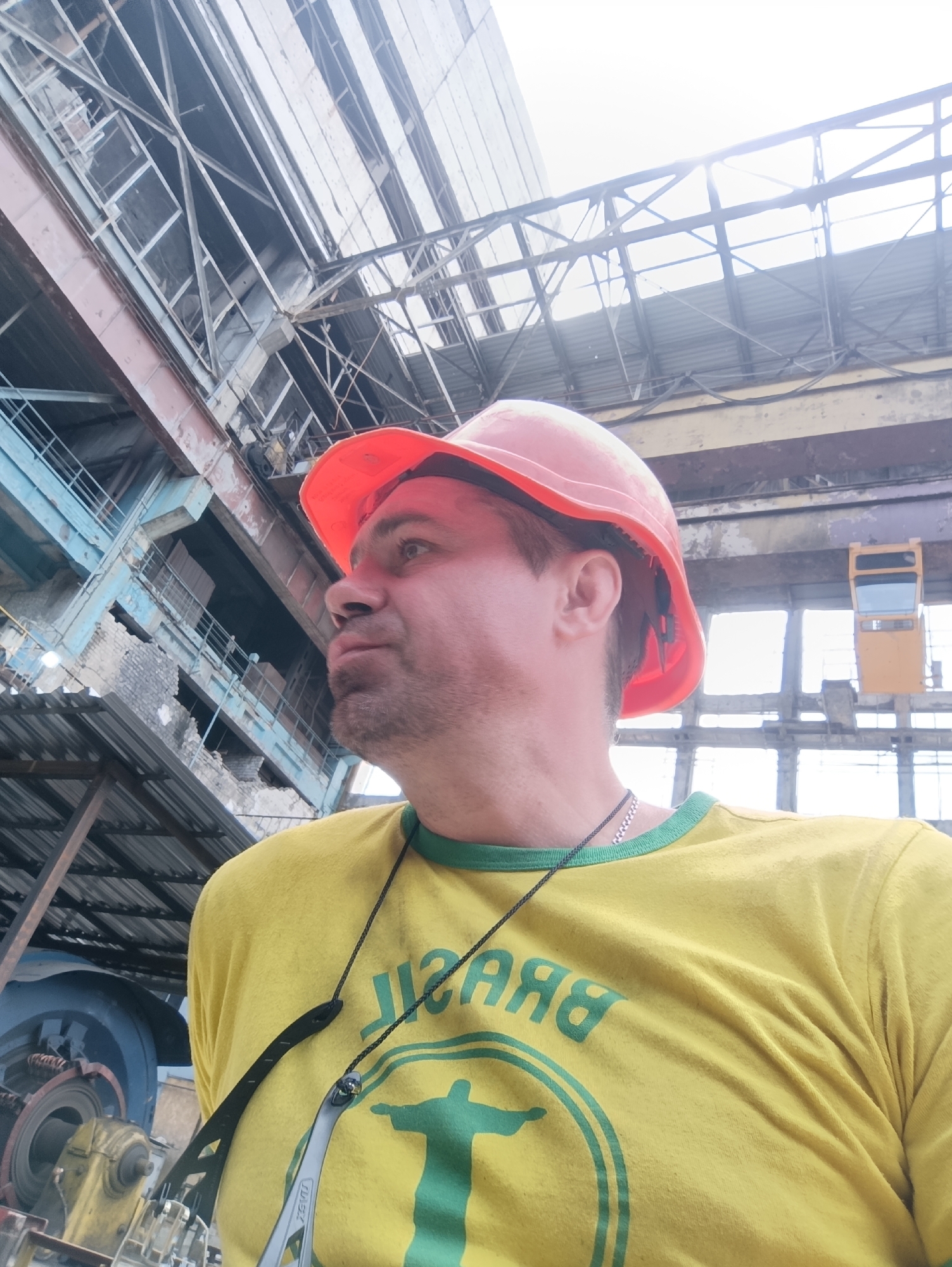 A man in an orange hard hat and a yellow t-shirt with a green design of "BRASIL" and a silhouette of Christ the Redeemer is shown from the chest up, looking to the left. He is wearing a silver chain necklace and a black strap is hanging around his neck. Behind him is a large industrial building with exposed metal beams and corrugated metal roofing. The lighting suggests it is daytime, with some sunlight filtering through. The overall environment appears to be an industrial or manufacturing facility, likely located within the city of Ladyzhyn, Ukraine, given the context.