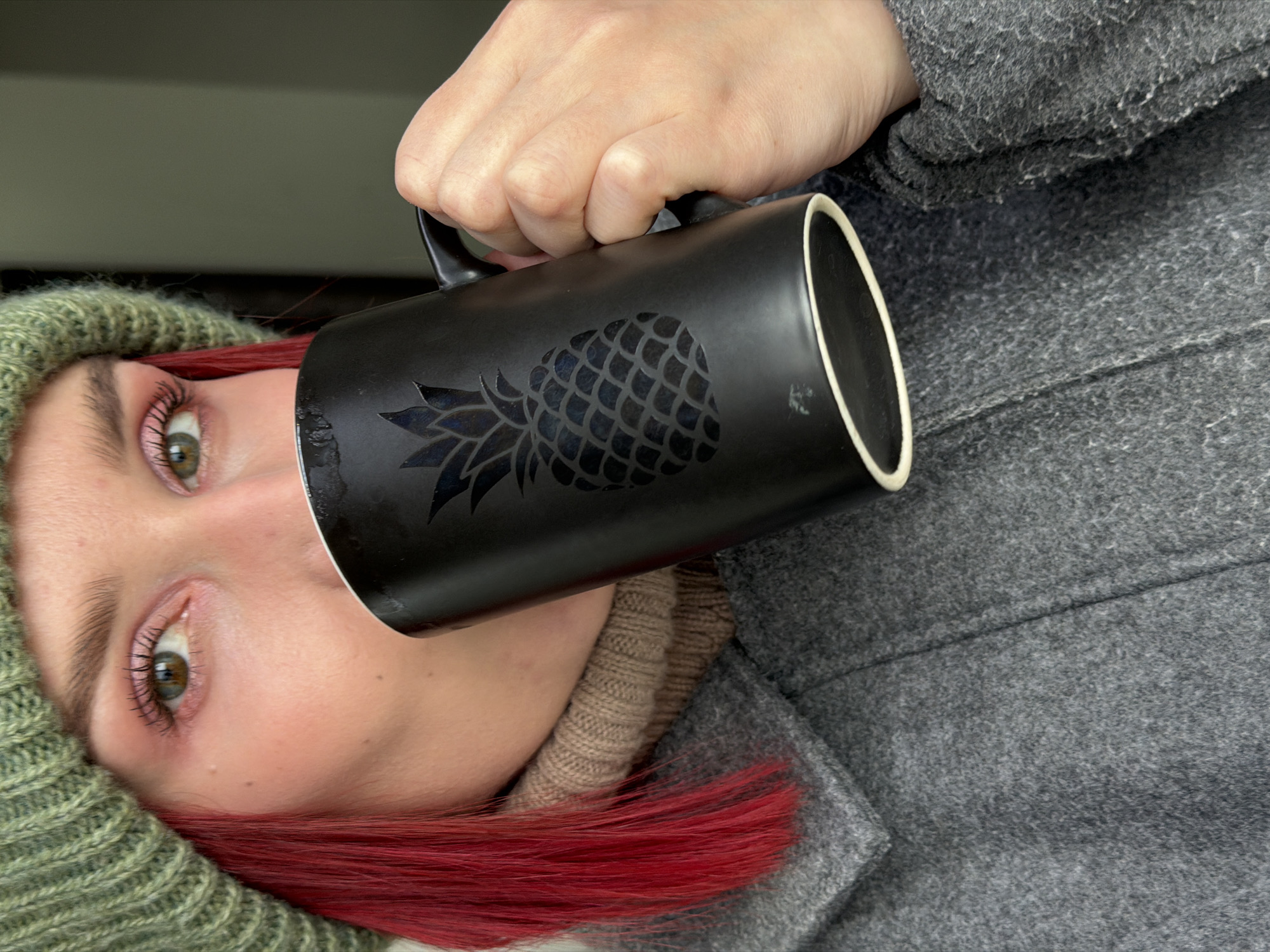 A person is depicted from the chest up, facing the camera with their head tilted slightly. They are holding a black mug with a cut-out or embossed pineapple design to their mouth, appearing to drink from it.
The subject has fair skin, distinct green or hazel eyes with visible mascara and subtle eye makeup, and light pink lips. They wear a knitted olive green beanie that partially covers their head, revealing vibrant red hair underneath. A beige knitted turtleneck or thick scarf is visible around their neck, layered beneath a textured, grey wool or felt coat.
The black mug is prominent, held by a hand with light skin tone. The inside of the mug shows a light-colored rim, possibly white or cream. The background is a soft, out-of-focus dark grey, suggesting an indoor setting. The warm clothing worn by the person implies a cool or cold environment, consistent with the location context of Chaikovsky, Russia. There is no visible text in the image, only the pineapple emblem on the mug. The overall impression is a casual, possibly cozy moment.