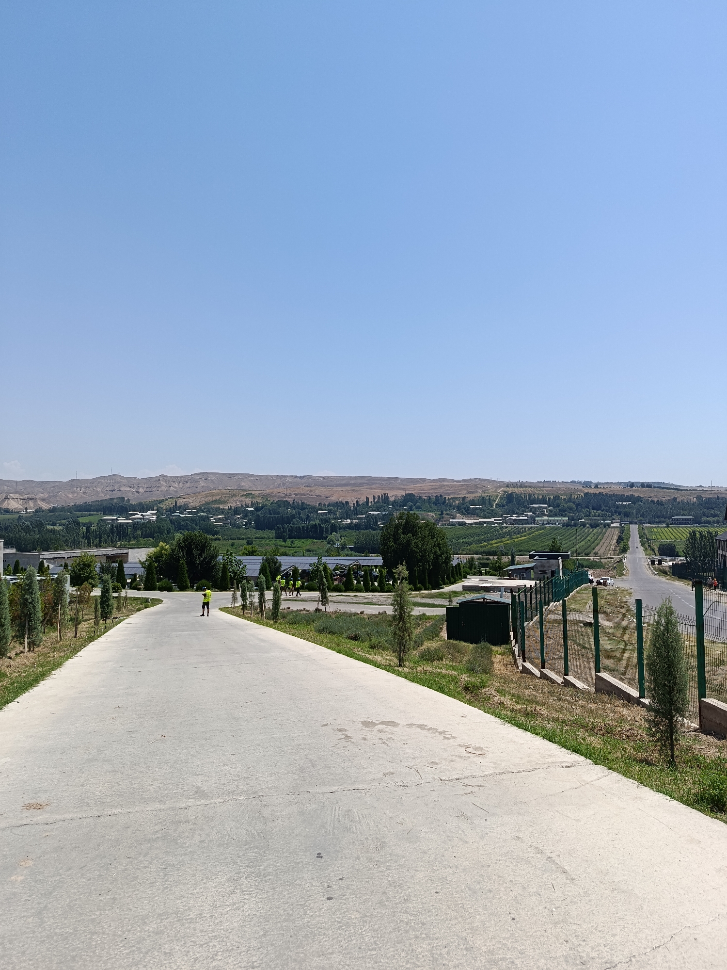 The image depicts an outdoor scene in Gulshan, Uzbekistan, under a clear blue sky. In the foreground, a wide, grey concrete path leads away from the viewer, bordered by short, manicured cypress trees on either side. A lone individual, wearing a bright yellow vest, stands in the middle of the path, facing away from the camera. Further down the path, a group of individuals, also wearing yellow vests, are visible, along with several vehicles. To the right of the path, a green fence encloses an area with sparse vegetation. Beyond the fence, a paved road curves into the distance, lined with trees and agricultural fields. In the background, a sprawling landscape of buildings, trees, and cultivated land stretches towards a range of dry, rolling hills. The overall impression is one of a semi-rural or developing area, possibly an industrial park or agricultural complex.