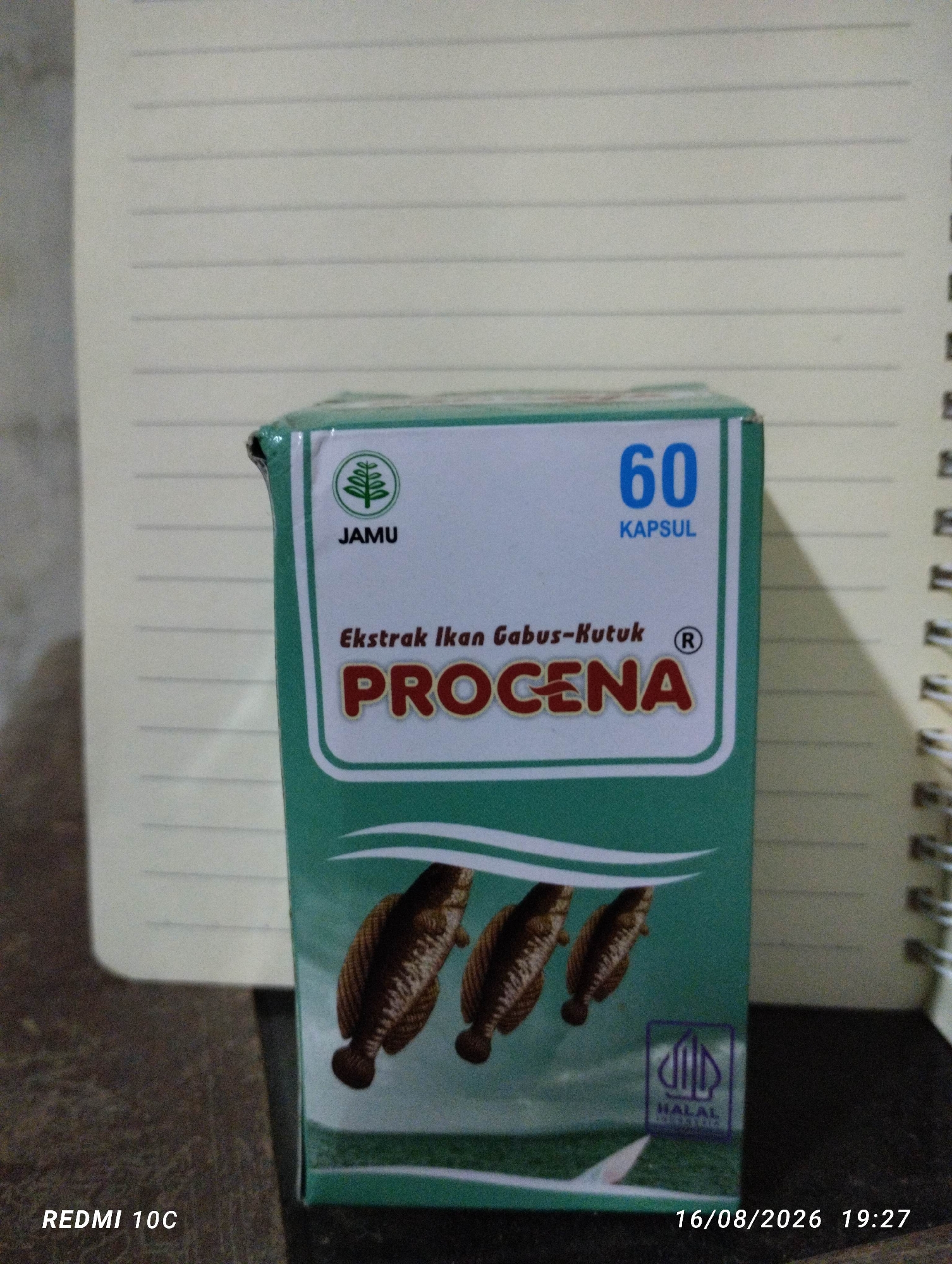The image shows a box of "PROCENA" capsules, a type of jamu (traditional Indonesian herbal medicine). The box is teal green with white lettering and graphics. It features the number "60" indicating the quantity of capsules, and the text "JAMU" above a green tree logo. Below the brand name, it states "Ekstrak Ikan Gabus-Kutuk" which translates to "Snakehead Fish Extract". Three fish are depicted on the front of the box, and a halal logo is visible in the bottom right corner. The box is placed on a surface that appears to be a notebook with lined paper, which is partially visible in the background. The bottom of the image contains a timestamp and the text "REDMI 10C", likely indicating the device used to take the photo. There are no people or specific locations within Kembiritan, Indonesia, visible in the image.