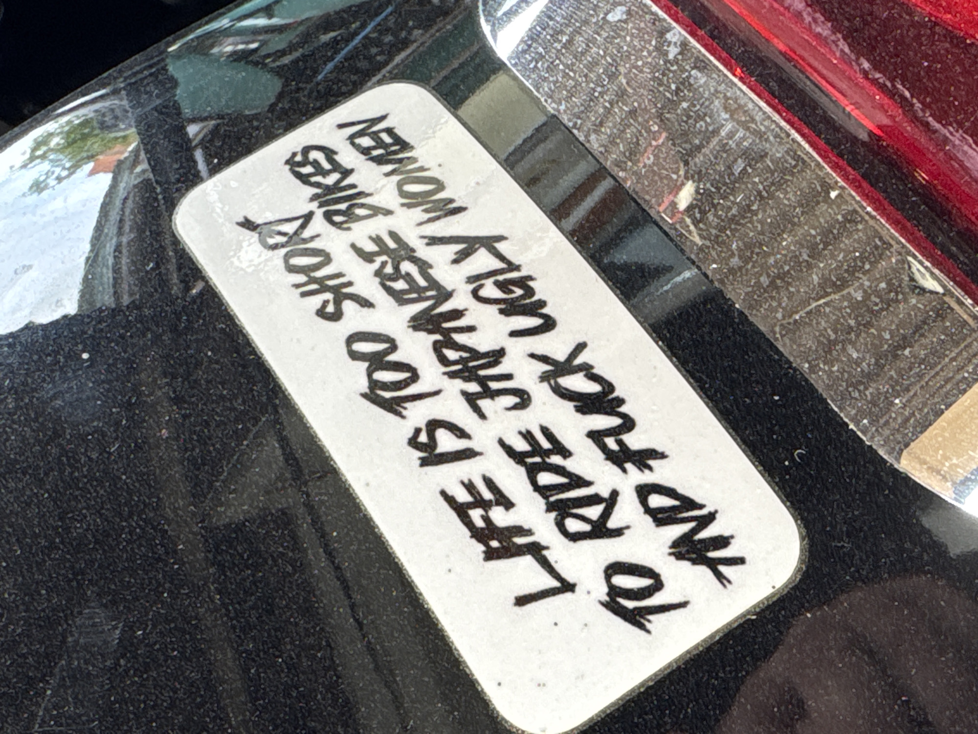 The image shows a sticker adhered to a dark surface, likely a motorcycle part based on the curvature. The sticker is rectangular with rounded corners and contains black text on a white background. The text is arranged vertically and reads: "LIFE IS TOO SHORT TO RIDE JAPANESE BIKES AND FUCK UGLY WOMEN". A portion of a red object, possibly a tail light, is visible in the upper right corner of the frame. The overall scene appears to be an outdoor setting based on the lighting. Location: The city of Krasnodar, Russia is not discernible from the visual information provided.