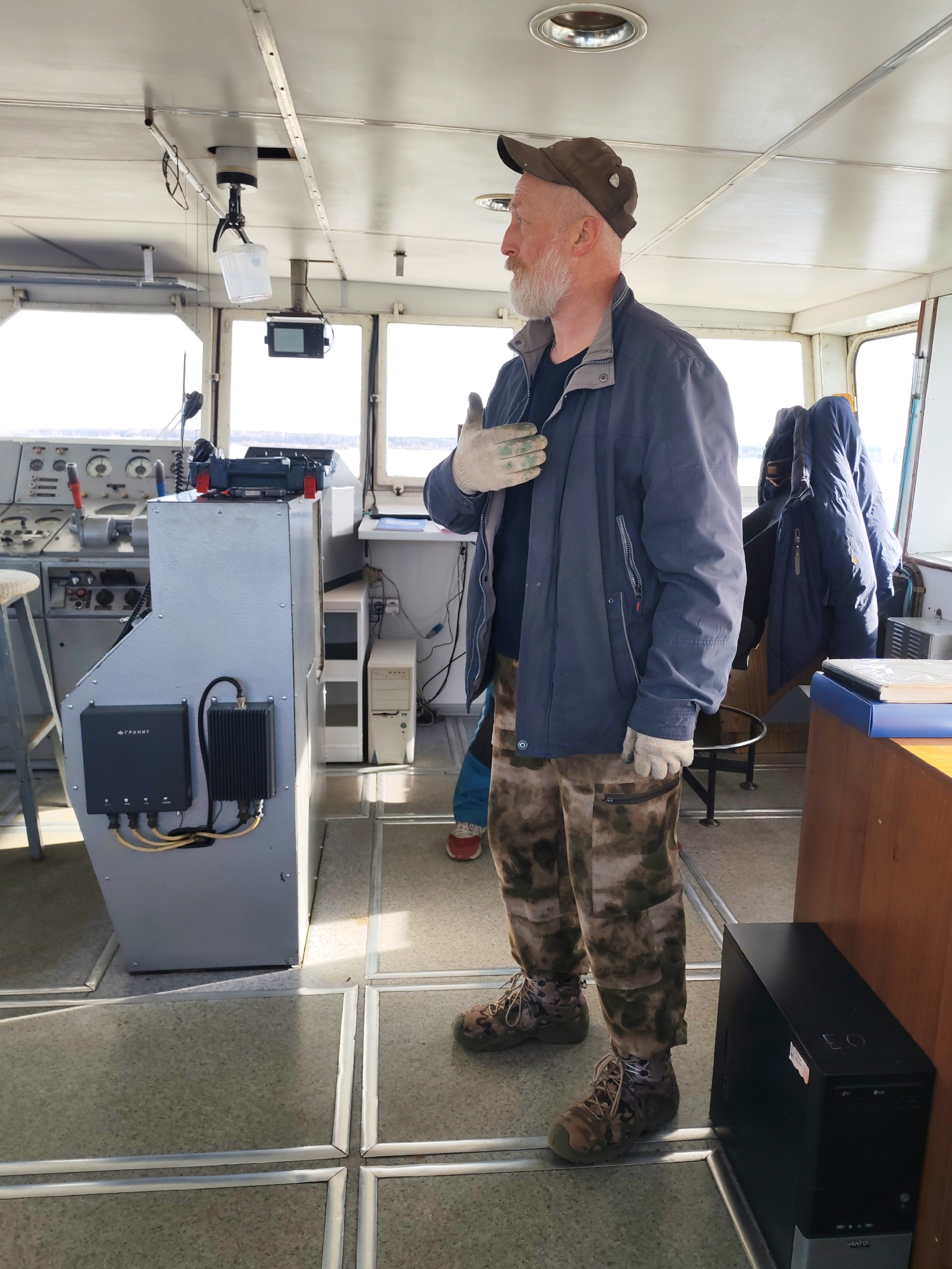 The image captures the interior of a boat or ship's pilothouse, centered on a bearded man standing and looking towards the right. The man, with a prominent white beard and wearing a brown baseball cap, a dark blue jacket, camouflage cargo pants, and light-colored work gloves, has a contemplative or observant expression. His left hand is raised to his chest.
The setting is clearly a functional marine vessel. To the left, a detailed control console with numerous gauges, levers, and communication devices is visible. Attached to the console's side is a grey enclosure featuring a black box labeled "ГРАНТ" (GRANT) and another ribbed black electronic unit, connected by yellow cables. A white computer tower stands behind the console. On the floor to the man's right, a black computer tower displays the "LG" logo and the text "VENTO" is partially visible.
The ceiling is light-colored and features integrated lighting. A distinctive detail is a white plastic bucket suspended from the ceiling by a gripper mechanism. Large windows offer a bright view of the exterior, appearing to be a wide body of water, possibly with ice or bright reflections, indicating clear daytime conditions. A glimpse of another person's leg and foot is seen behind the console on the left. Several dark coats are hanging on a rack in the background on the right. The floor has a grid-like pattern of grey tiles separated by metallic strips. The overall scene suggests a routine operation or observation within the command center of a vessel.