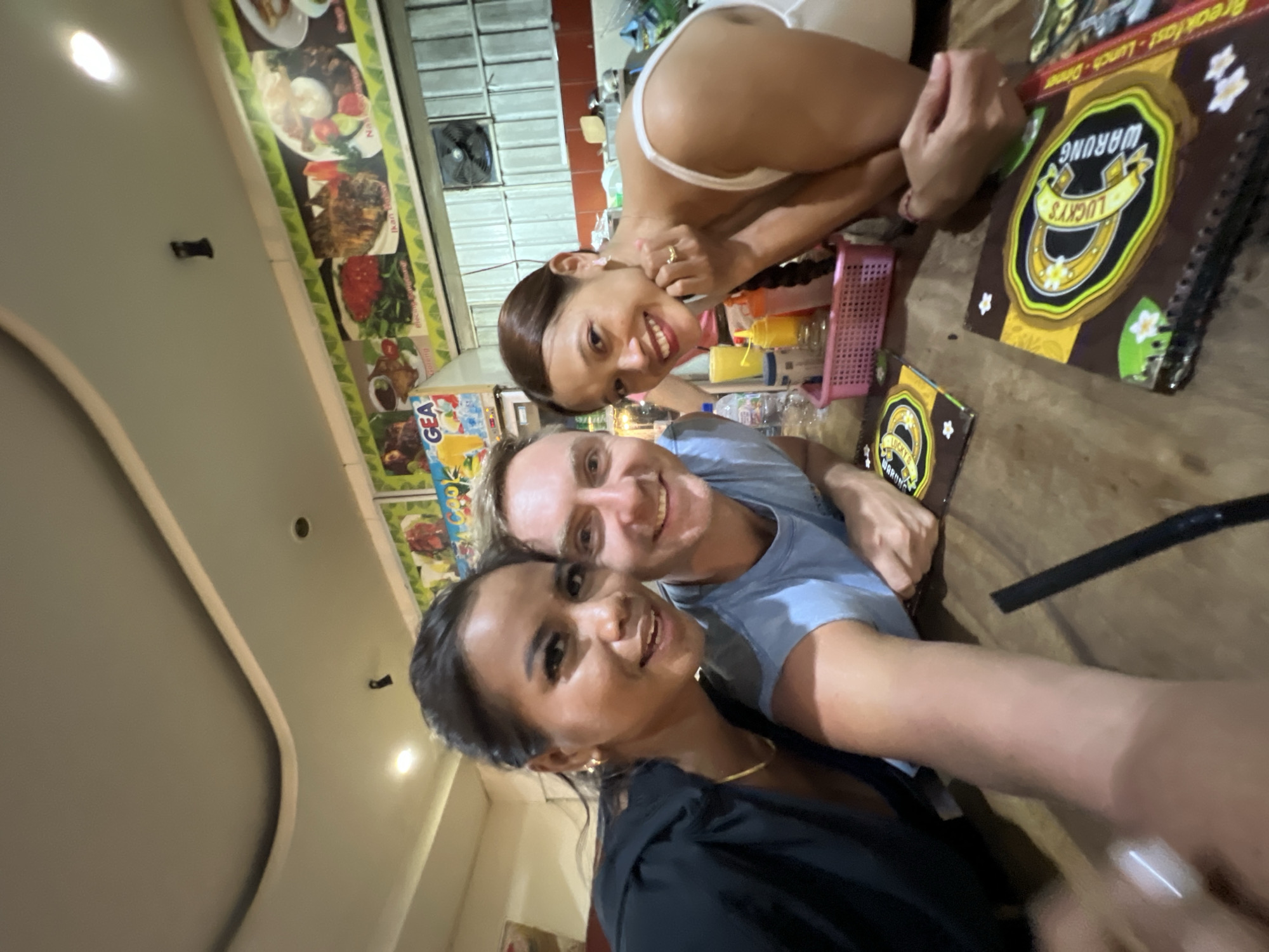 The image shows three people taking a selfie at a restaurant in Kuta, Indonesia. The foreground includes a female wearing a black top and gold necklace. Behind her is a man wearing a blue shirt. To the side of the man is another woman wearing a white top and gold ring. On the wooden table are two menus for "Lucky's WARUNG" and several plastic bottles. Behind the group is a wall with images of different meals and an air conditioning unit.