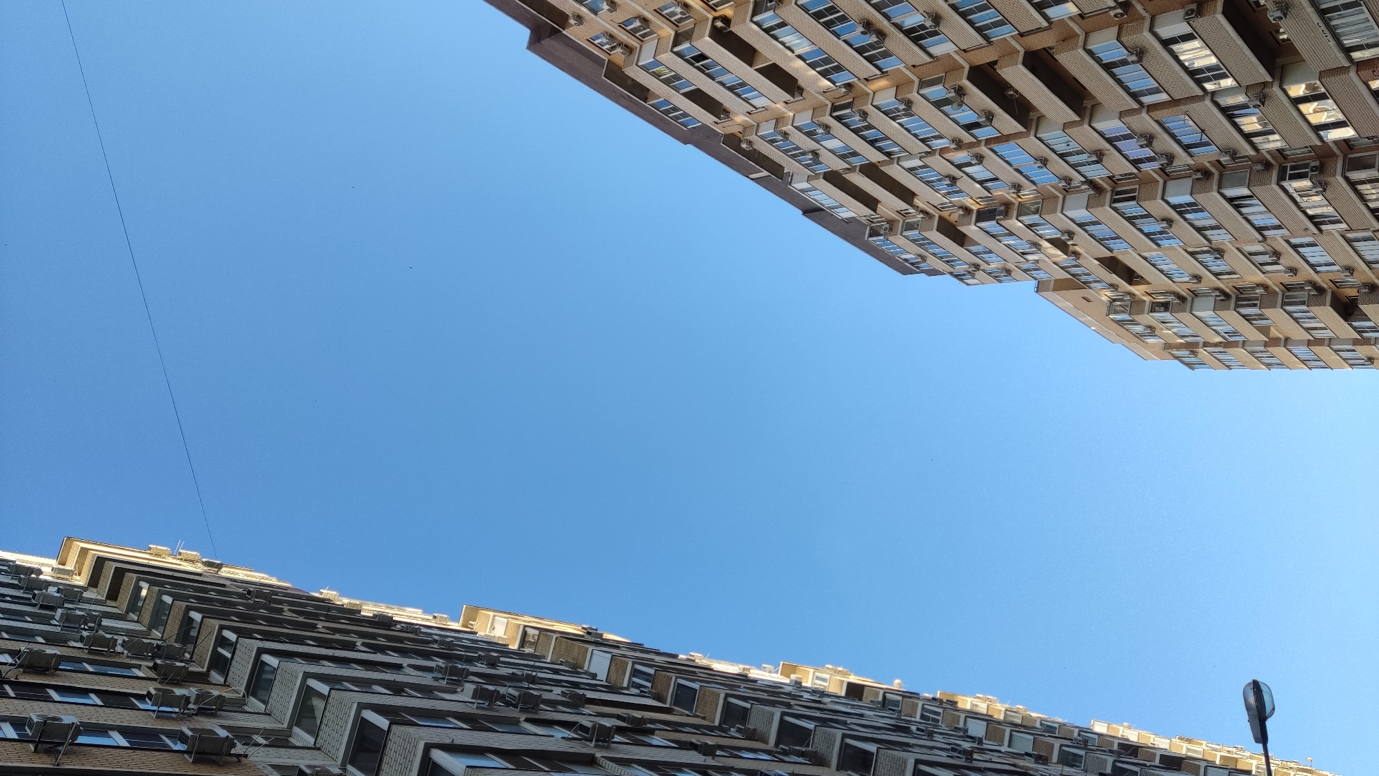 This is a low-angle shot of two apartment buildings against a clear blue sky. The building on the left is to the foreground and angled towards the camera, showing a facade covered in windows and air conditioning units. The building on the right is further away and also angled towards the camera, displaying a similar architectural style with multiple stories, balconies, and numerous windows. A street lamp is visible in the lower-left corner. The image appears to be taken in Krasnodar, Russia, as indicated by the typical Soviet-era architecture of the apartment buildings. There are no people visible in the frame.