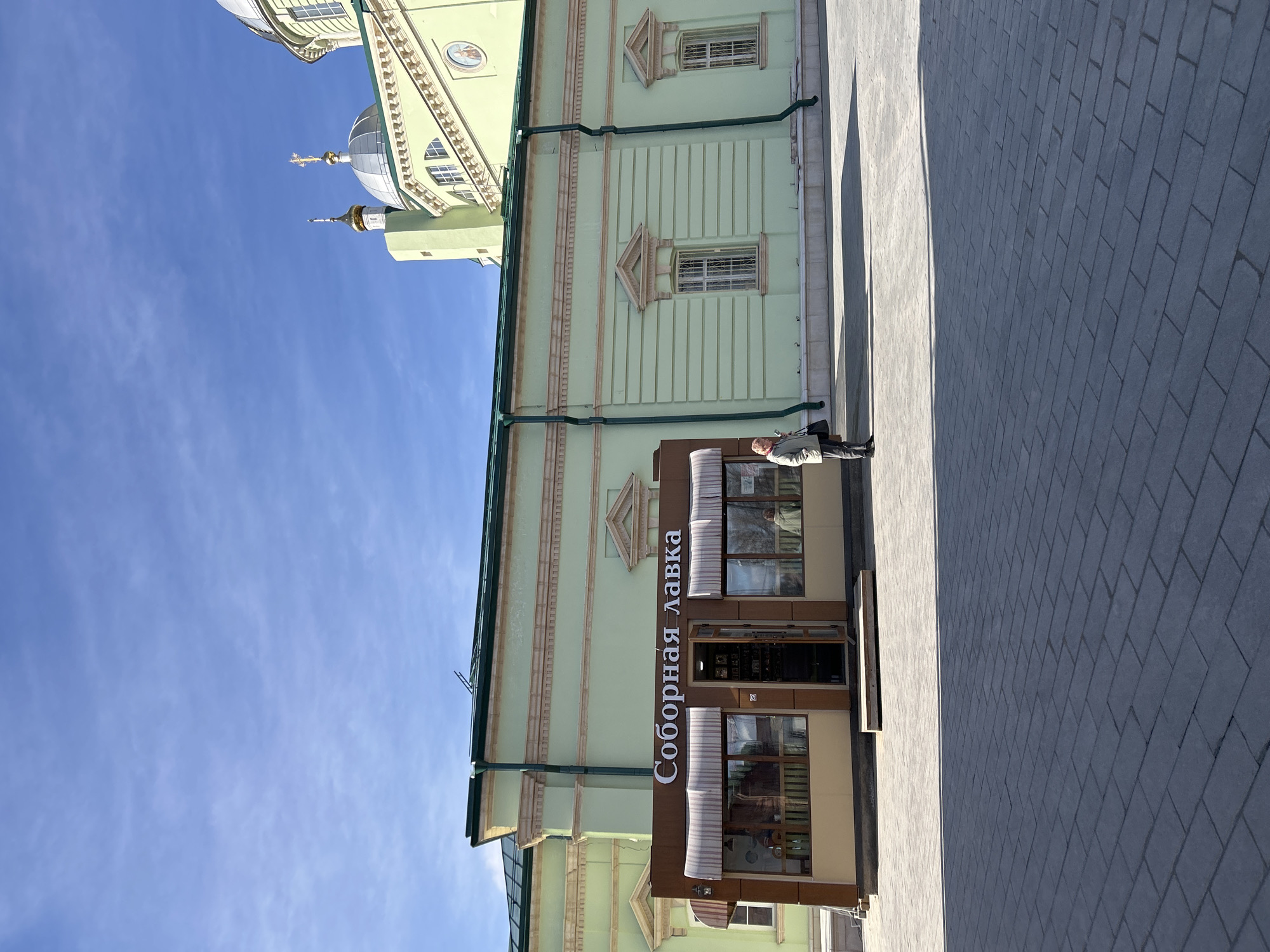 A vertical, wide-angle photograph shows a building in Klin, Russia, under a clear blue sky. A portion of the building's exterior is light green with beige trim and architectural details such as triangular window accents. A sign vertically reading "Соборная Лавка" (Sobornaya Lavka) is attached to the building, above a storefront with a brown frame. A person stands near the entrance. The sidewalk in the foreground is paved with grey rectangular stones. In the upper-left, part of a church with a golden dome is visible.