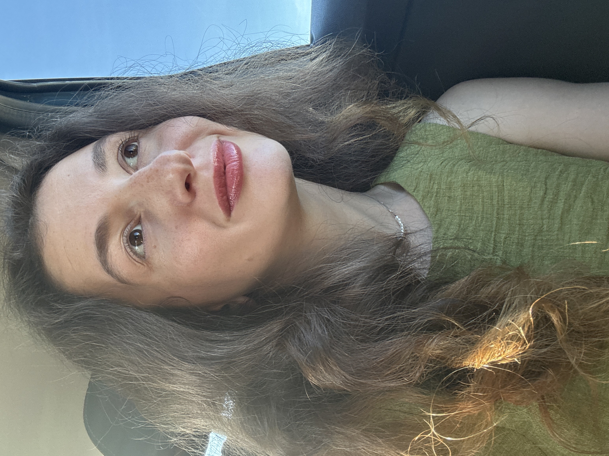 A medium shot captures a young woman from the chest up, looking to her right with a slight smile. She has long, wavy, dark brown hair that frames her face. Her skin has a warm tone with visible freckles. She is wearing a textured, sleeveless, green top and a delicate silver necklace. The background is divided between a bright blue sky and a dark interior, suggesting she is in a vehicle. The lighting appears to be natural, coming from her right, highlighting strands of her hair.