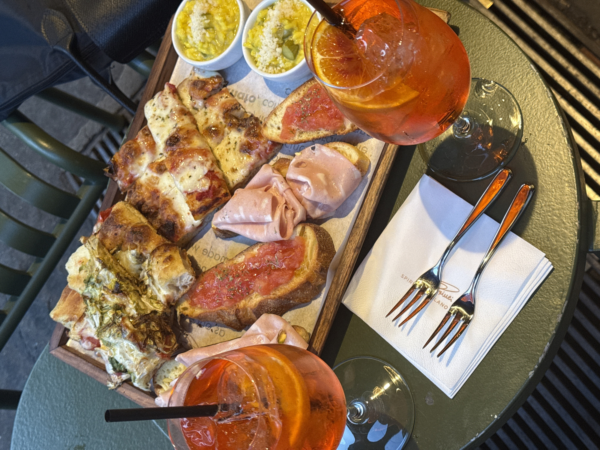 This image displays an aperitivo spread on a dark green, textured outdoor table in Milan, Italy. The central feature is a wooden serving tray containing various food items.
On the table, two stemmed glasses hold an orange-red beverage, consistent with an Aperol Spritz, each containing ice cubes, an orange slice, and a black straw. The wooden tray, lined with paper featuring faint, repeating text, presents an assortment of appetizers: multiple rectangular slices of focaccia-style bread, some topped with melted cheese and herbs, others with a cooked green leafy vegetable; several slices of toasted bread topped with a red, tomato-based spread and dried herbs; and folded slices of light pink cured meat.
Two small white ceramic bowls, situated at the top right of the tray, contain a yellow, granular dish, garnished with grated white cheese and small green pieces. A stack of white paper napkins, one featuring partially visible text "SPIRITO MILANO," and two silver forks with distinctive orange handles are positioned on the table in the foreground. The background is blurred, revealing dark green metal outdoor chairs with horizontal slats and a dark grate beneath the table. No individuals are visible.
