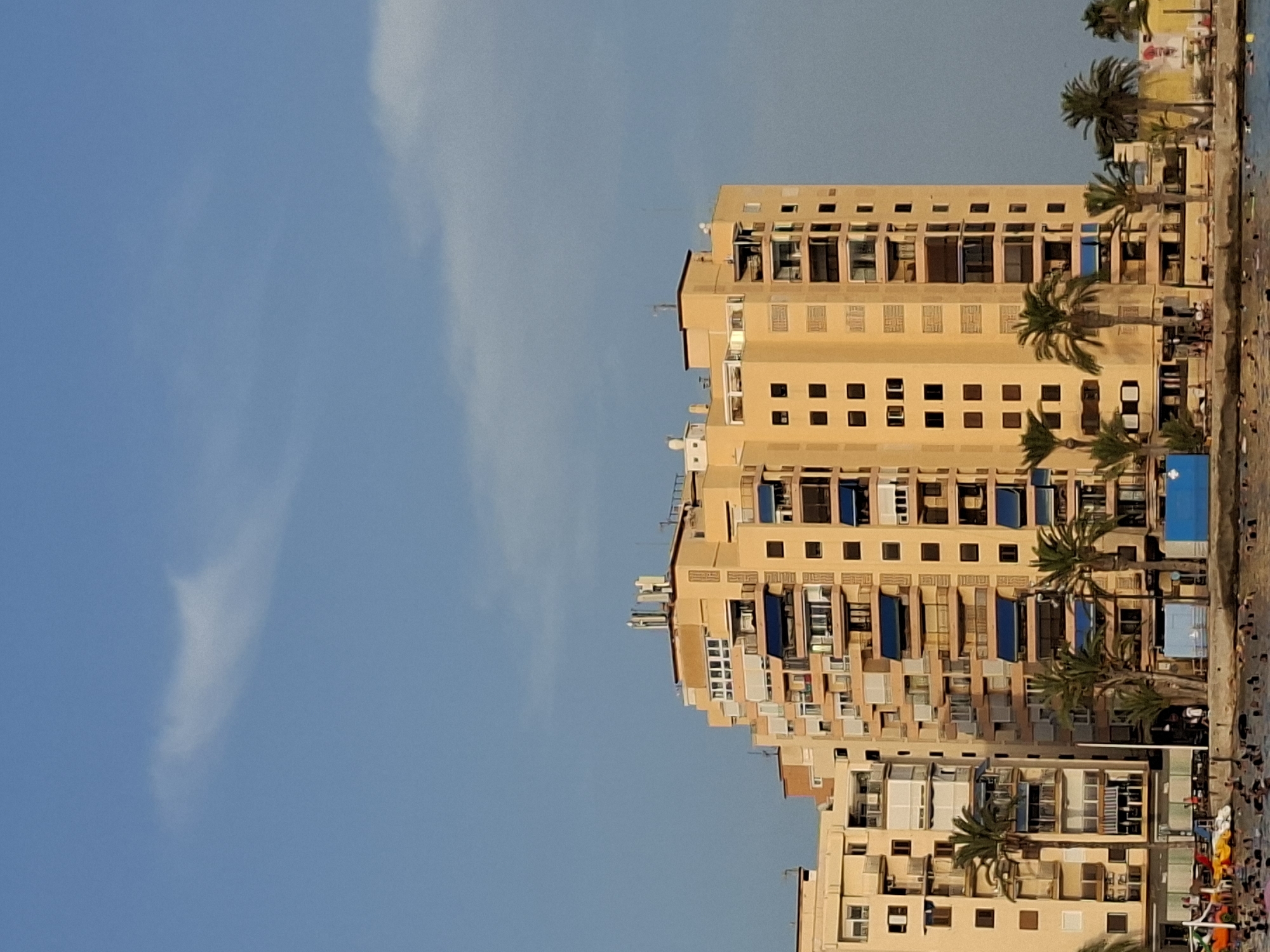 The image shows a seaside scene in Torrevieja, Spain, featuring a tall, yellow apartment building with many balconies and blue awnings. The building is situated next to a body of water, likely the Mediterranean Sea, where numerous people are swimming and several small boats are visible. Palm trees are scattered around the base of the building and along the shoreline, and a clear blue sky with a few wispy clouds forms the backdrop. The architecture of the building suggests a coastal resort or residential area.