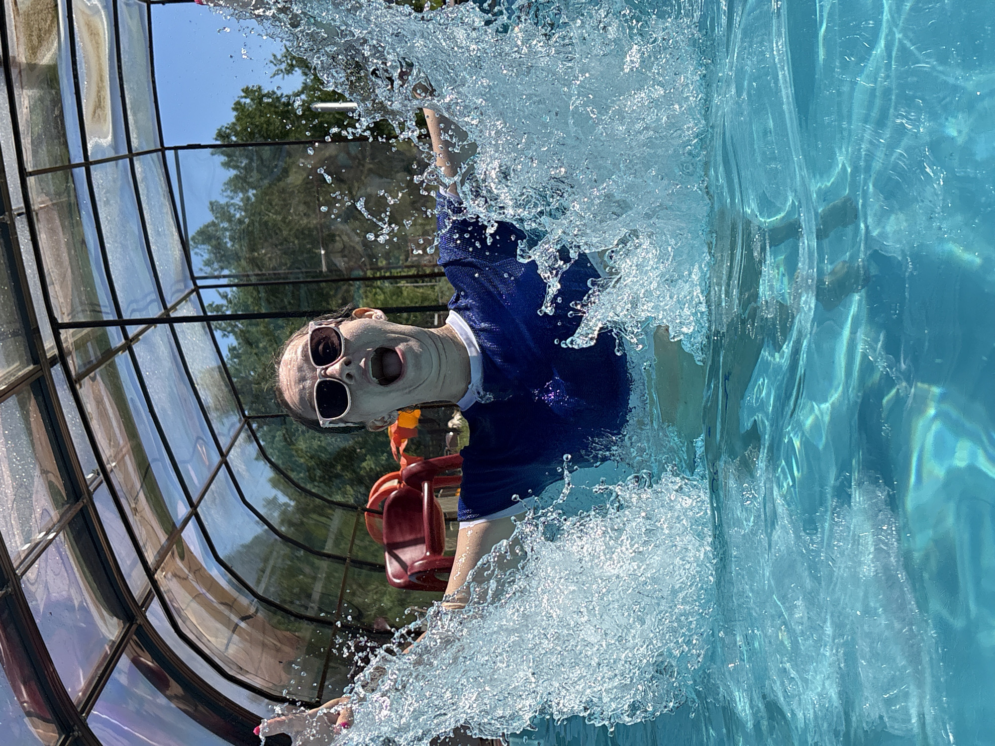 A young person with wide eyes and an open mouth is splashing in an indoor swimming pool. They are wearing a blue t-shirt with a white collar and white sunglasses. The water is splashing dramatically around them, with droplets visible in the air. The pool is covered by a transparent, curved roof structure made of metal and plastic. Outside the pool enclosure, trees and a bright blue sky are visible. A red chair is situated behind the person in the pool. This scene is taking place in the city of Birobidzhan, Russia.