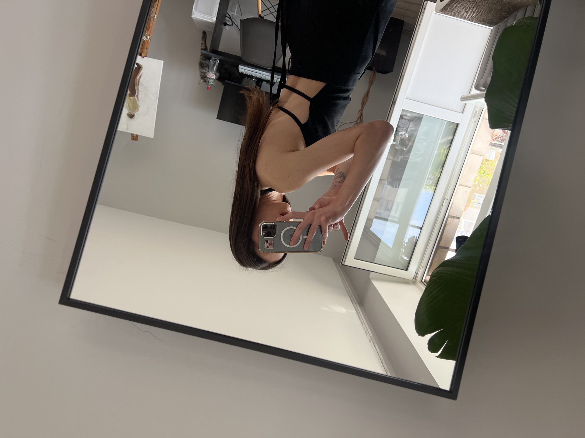 A woman in a black outfit is taking a selfie in a mirror. The mirror reflects a room with a window, a desk with various objects, and a plant. The location cannot be identified from the image.