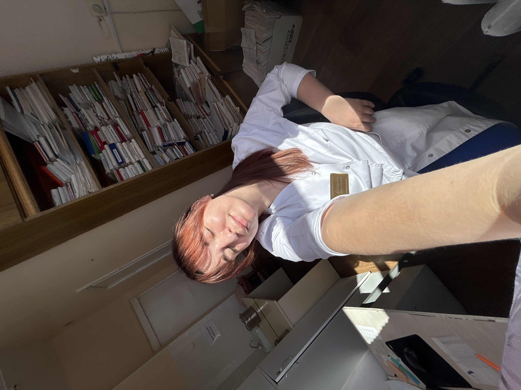 A woman with auburn hair is lying on a chair, likely in an office setting in Фоки, Russia. She is wearing a white lab coat, and there is a badge pinned to the coat. A filing cabinet is visible to the left, filled with paper documents. The environment appears to be an organized but cluttered workspace, potentially a medical office or similar professional environment. Additional furniture or equipment can be seen in the background, but they are not in clear focus.