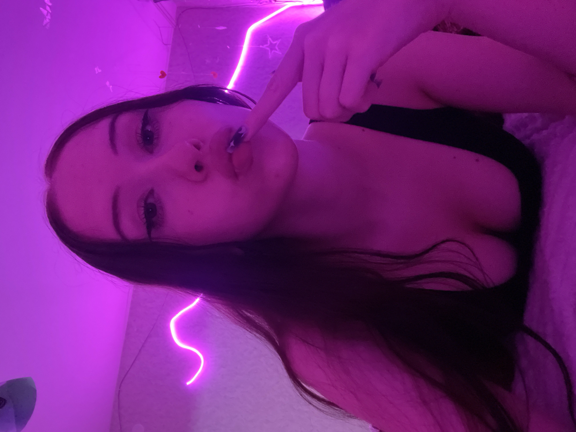 A young woman with long dark hair and full lips is shown in a close-up shot. She is wearing a black top and has her finger gently placed on her lips. The scene is illuminated by pink neon lights, which create a vibrant and moody atmosphere. The background appears to be a bedroom, with hints of a wall and ceiling visible, decorated with pink neon light strips in a wave pattern and some small heart shapes. The overall impression is one of intimacy and personal expression, with the neon lighting adding a modern and aesthetic touch. The image does not contain information about the location in Votkinsk, Russia.