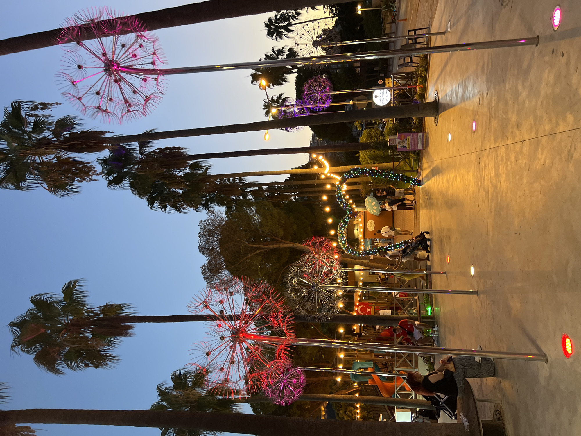 The image depicts an outdoor scene at dusk in Alanya, Turkey, likely a public walkway or park area. Several large, illuminated dandelion-shaped sculptures, with pink and red lights, are prominent features. Palm trees are scattered throughout the scene, some with their fronds illuminated by the ambient light.
In the foreground, a woman with long red hair is seated on a wooden bench, facing to the left. She is wearing a black top and patterned trousers. To her right, a man in a red t-shirt and white shorts is also seated on the bench. A child is visible behind them. The ground is paved with concrete and features several colored lights embedded in it, casting glows of red and pink.
Further into the scene, there is a children's playground area with colorful slides and structures. People are walking around, some pushing strollers. String lights are hung overhead, and a large arch decorated with colorful lights forms an entrance or walkway. A Turkish flag is visible on a pole behind the playground.
In the background, buildings and more palm trees are silhouetted against the twilight sky. The overall atmosphere suggests a lively, festive evening setting.