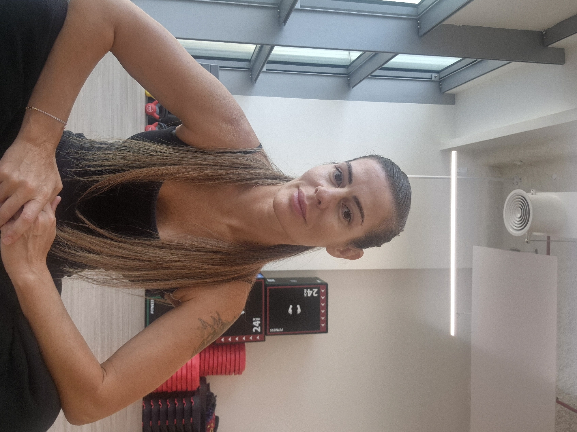 The image depicts a woman in a fitness setting. She has long, straight brown hair and is wearing a black tank top. There is a visible tattoo on her upper arm. Gym equipment, including a black box labeled "24 INCH FITNESS," and dumbbells are located in the background. The room is well-lit, possibly due to the overhead skylight structure visible in the upper portion of the frame. A light fixture and ventilation system are mounted on the wall in the background. The location is Unknown, Unknown.