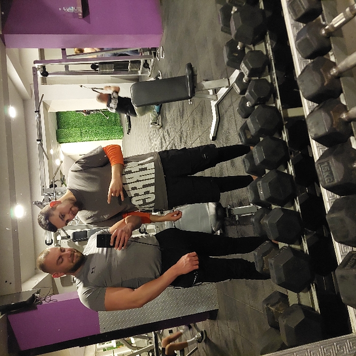 The image depicts a mirror selfie taken inside a gym facility in Pazardzhik, Bulgaria. Two adult males are prominently featured in the foreground, reflected in a large mirror. The male on the left, bald with a beard, is wearing a grey short-sleeved athletic shirt and dark pants; he holds a black smartphone horizontally, aimed towards himself and his companion, indicating he is taking the photograph. His left arm is flexed. The male on the right, with short dark hair and a beard, is wearing a grey t-shirt with partially visible "PERFECT" text over an orange long-sleeved shirt, black shorts, and black leggings; he is looking towards the phone and making a "rock on" hand gesture with his right hand.
The lower portion of the reflection shows a metal dumbbell rack stocked with numerous black hexagonal dumbbells, some labeled "15KG." The gym interior, visible in the background reflection, features dark rubber floor mats, a purple painted wall section on the right with a small shelf holding a water bottle, and a green artificial turf-textured wall panel in the center-left. Various gym equipment, including cable machines and an adjustable utility bench, are visible. Multiple other individuals are present: one male is seen kneeling and operating a cable machine in the mid-right background, another person is partially visible near equipment in the far right, and the lower legs and feet of a person are visible in the far left background. Overhead artificial lighting illuminates the scene.