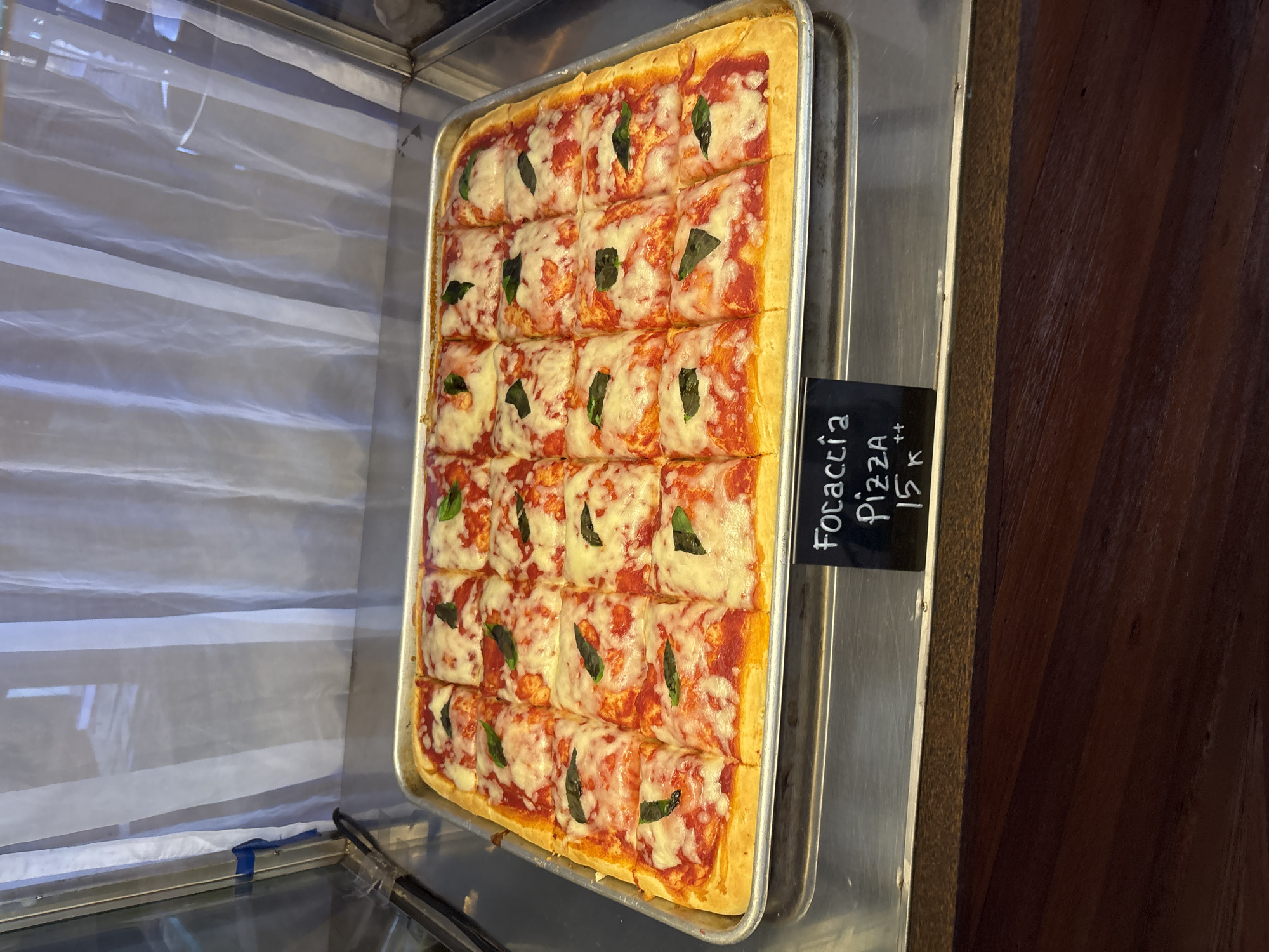 The image shows a tray of focaccia pizza, cut into individual square portions, displayed within a refrigerated display case. Each piece is topped with tomato sauce, melted cheese, and a single basil leaf. A small black sign indicates that the pizza is focaccia and priced at 15k ++ (likely referring to 15,000 Indonesian Rupiah plus taxes). The location is Pecatu, Indonesia. The tray sits on a stainless steel surface. A striped translucent plastic screen or barrier is visible in the background within the display case. The right side of the image shows a dark wooden surface, likely part of the display unit or surrounding furniture.