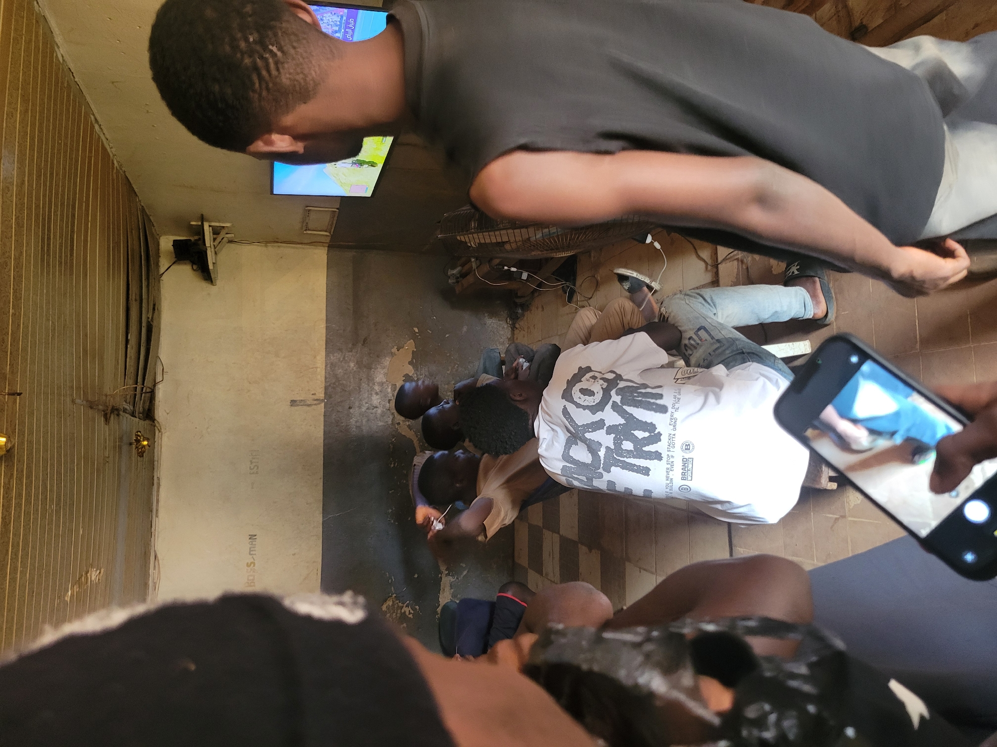 In Bokkos, Nigeria, a group of young men are gathered in what appears to be a common room or lounge area, likely a home or community space. They are engrossed in watching television, with several individuals seated and lying on the floor facing a TV screen mounted on the wall. The room has a rustic and somewhat unkempt feel, with exposed wiring, corrugated metal sheeting on one wall, and a visible fan. The lighting suggests it is daytime, though the interior is somewhat dim.
One man in the foreground is in profile, wearing a dark t-shirt, and his head is tilted upwards as if looking at the TV or something beyond the frame. Behind him, several other men are positioned on the floor, some lying down, others sitting up. One man is wearing a white t-shirt with a graphic design. Another is seen wearing a light-colored t-shirt. The overall atmosphere seems relaxed and communal, typical of friends or family spending time together.
In the lower right corner, a hand holding a smartphone is visible, capturing the scene. The phone screen displays a partial image of someone's legs and feet. The text "BOSS-MAN" and "ESTI" are visible on the wall in the background. The text "طيران الرياض" (which translates to "Riyadh Air") is seen on the television screen, suggesting a sports broadcast or news program. The image conveys a sense of casual leisure and shared experience.