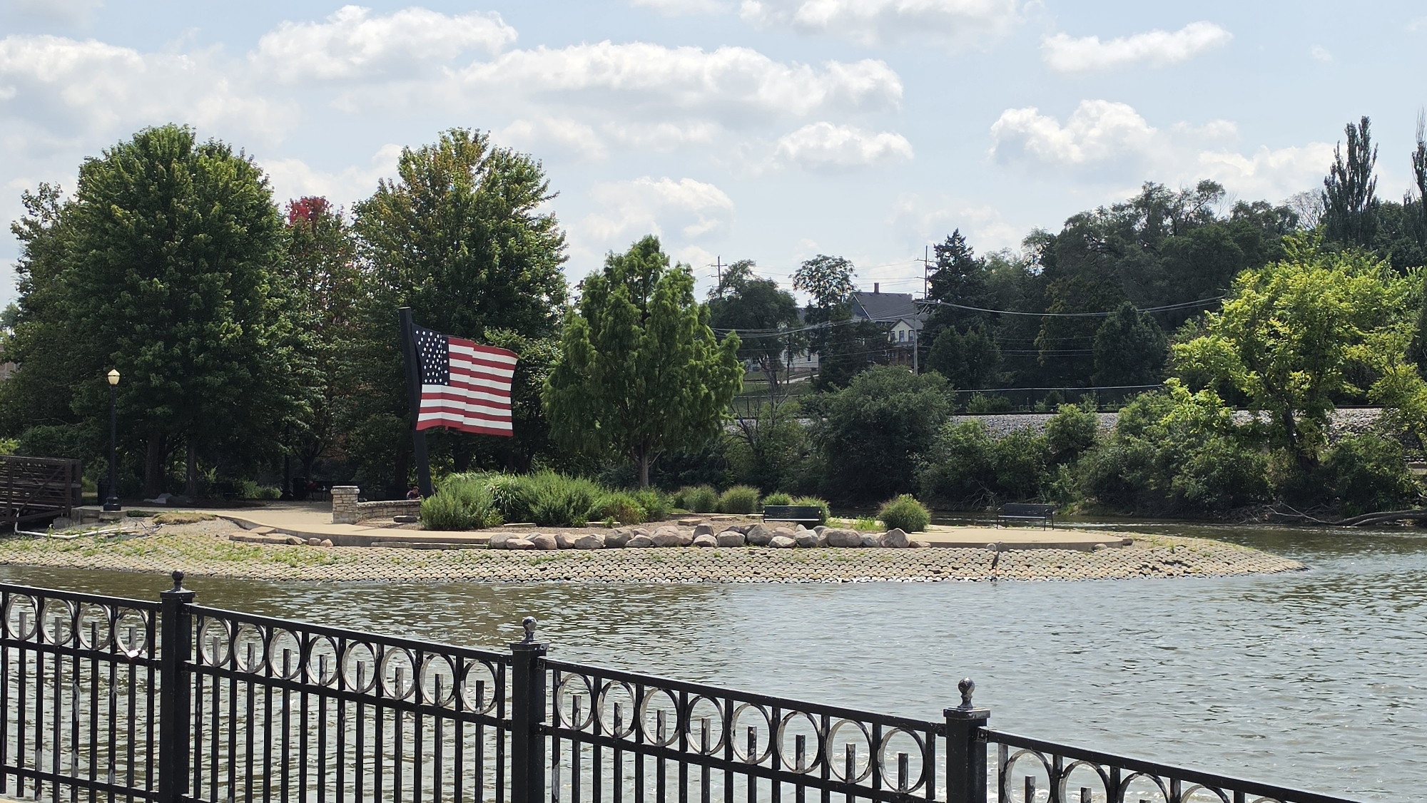 An American flag waves from a flagpole on a small island in the Fox River in Elgin, Illinois. The island is landscaped with grass, bushes, and large rocks. A black metal fence is in the foreground, obscuring some of the water. Behind the island, trees and houses are visible on a hill. The sky is partly cloudy.