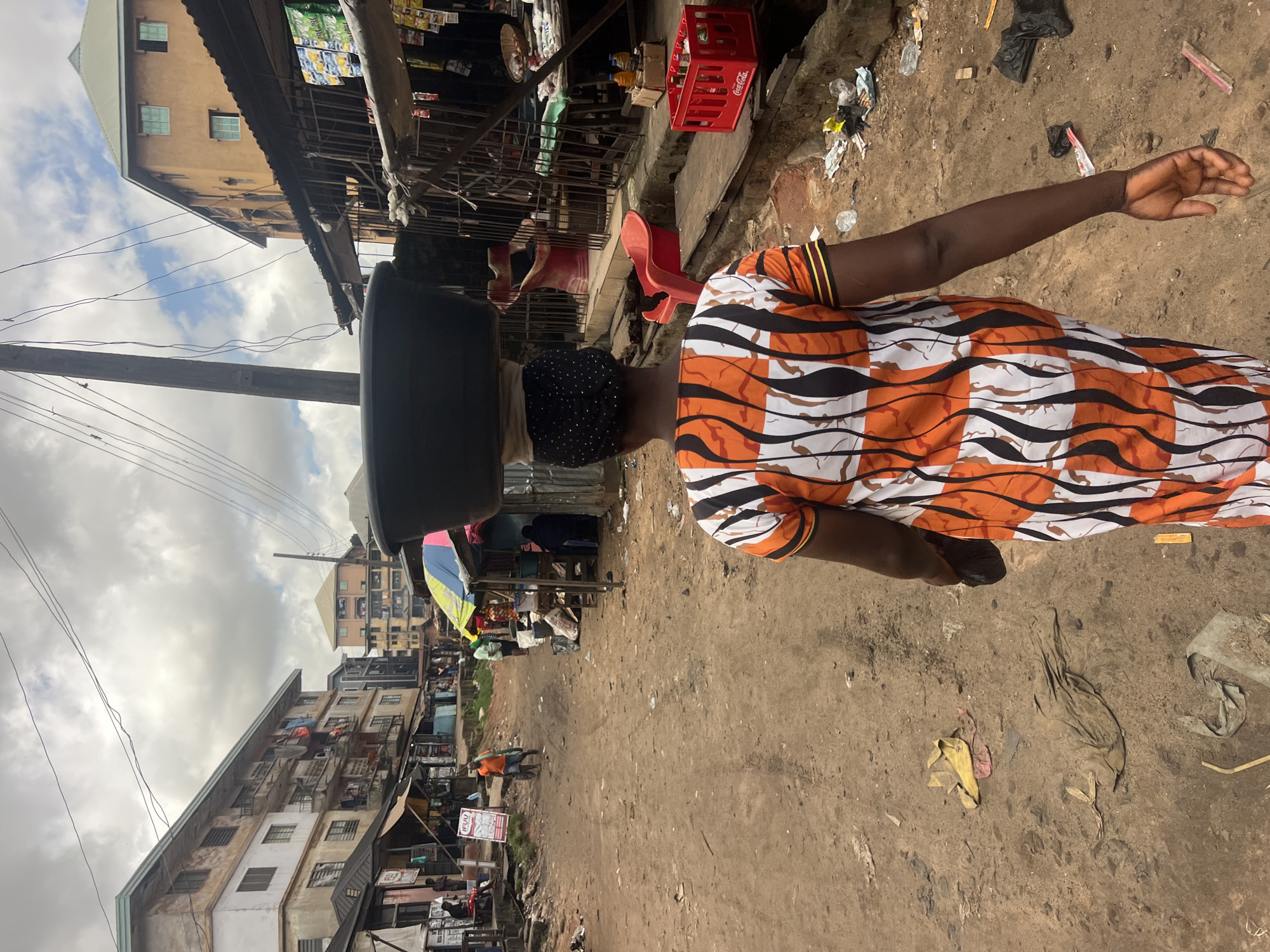 A woman walks away from the camera in a dirt street in Nkpor, Nigeria. She is wearing a long, colorful dress with an orange, black, and white abstract pattern and has a large black bowl balanced on her head. She is carrying a dark object in her right hand. The street is lined with shops and apartment buildings. The sky is cloudy. Debris litters the ground.