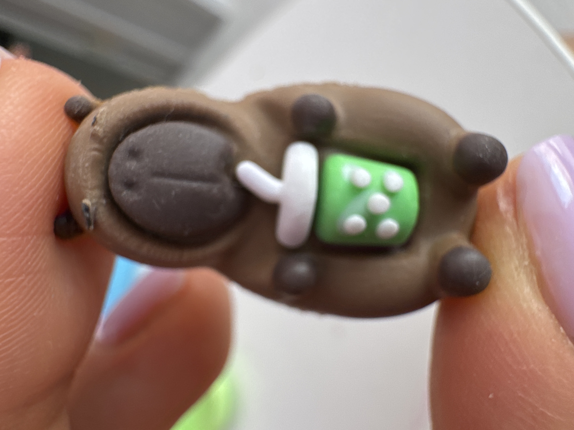A macro photograph shows a small, brown, inanimate figurine of a capybara wearing a hoodie and holding a green cup with a white straw. The figurine has a dark brown nose and two small, dark brown ears. The cup has white dots on its exterior. The figurine is held between two human fingers, one on the top with visible fingerprints and the other on the bottom. The background is blurred and appears to be a white surface with hints of blue and green. The image is an extreme close-up, with the figurine taking up most of the frame. No location in Uyutnoye, Ukraine, is visible.