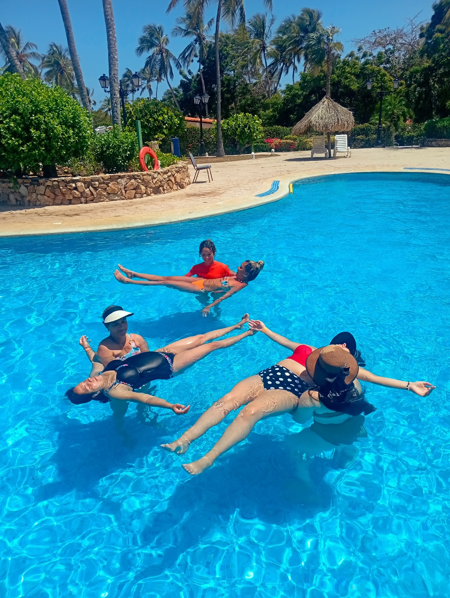 Five people are floating in a swimming pool in Playa El Agua, Venezuela. The pool is surrounded by palm trees, plants, and a stone wall. A lifeguard ring is visible on the side of the pool. Four of the people are lying on their backs, floating with their arms outstretched. One person is holding another person in the air. The person being held is lying horizontally across the arms of the other. The people are wearing swimwear of varying designs. One person wears a wide-brimmed hat. The water is clear and blue.