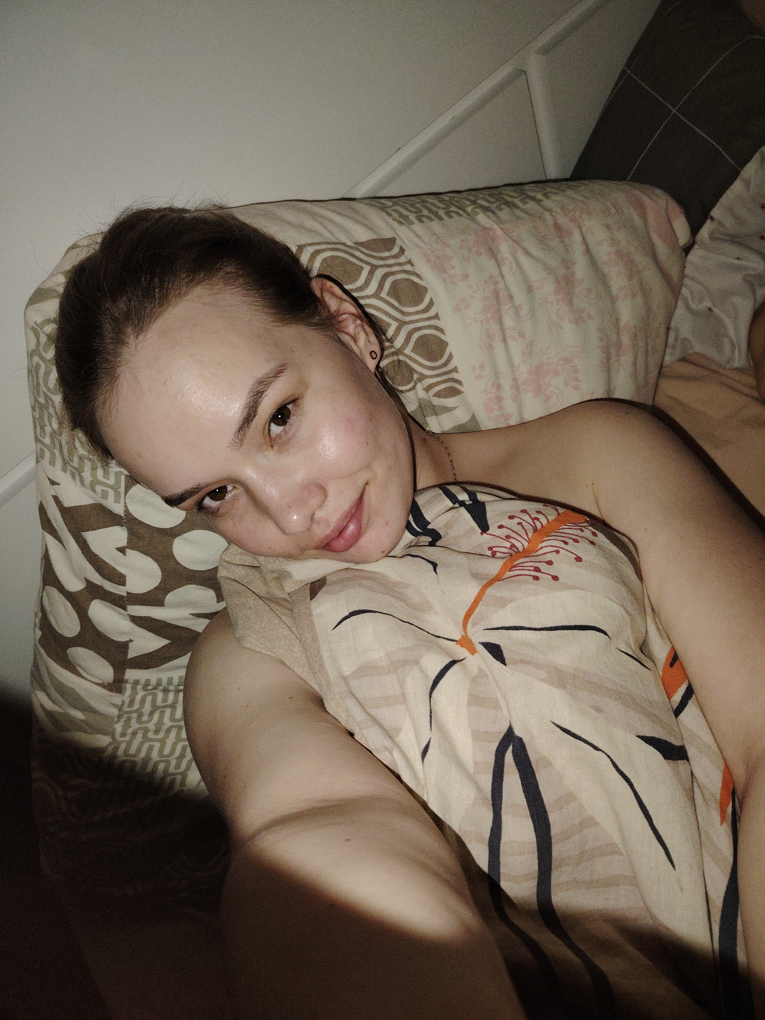 The image depicts an indoor scene in the city of Марковский, Russia. One individual, appearing to be an adult female with fair skin and dark brown hair, is lying on their left side in a bed. Their head is resting on a pillow with a brown and off-white geometric pattern. The individual is looking directly towards the camera, exhibiting a slight smile. A small dark stud earring is visible in the left earlobe, and a thin gold-colored chain is around the neck. The upper torso is partially covered by a duvet or blanket with an off-white background patterned with thin black lines and scattered orange, stylized floral elements. A light-colored garment is visible on the individual's right shoulder. Behind the individual, a second pillow displays a light pink floral pattern on an off-white ground. A dark gray cushion with a subtle grid pattern is partially visible further back. A white, possibly slatted, headboard or furniture element is against a plain light-colored wall. The scene is dimly lit, with a distinct shadow occupying the lower left portion of the frame.