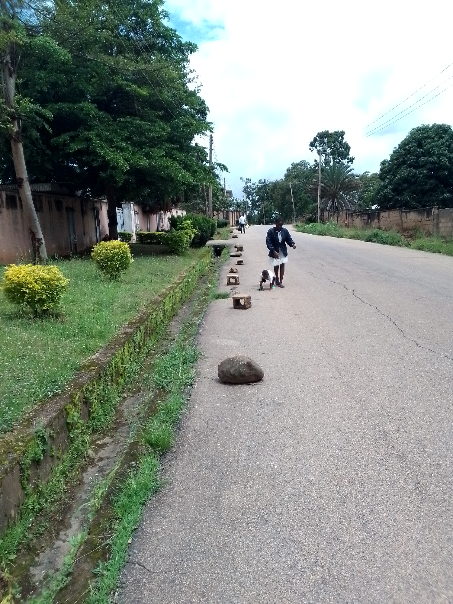 The image shows a paved road in Jos, Nigeria, with a grassy verge and a drainage channel on the left. Several rounded stones and rectangular blocks are placed along the edge of the road. A woman wearing a dark jacket and white skirt is walking on the road with a young child who is crawling on the ground in front of her. Trees and buildings line the street in the background. The sky is cloudy.