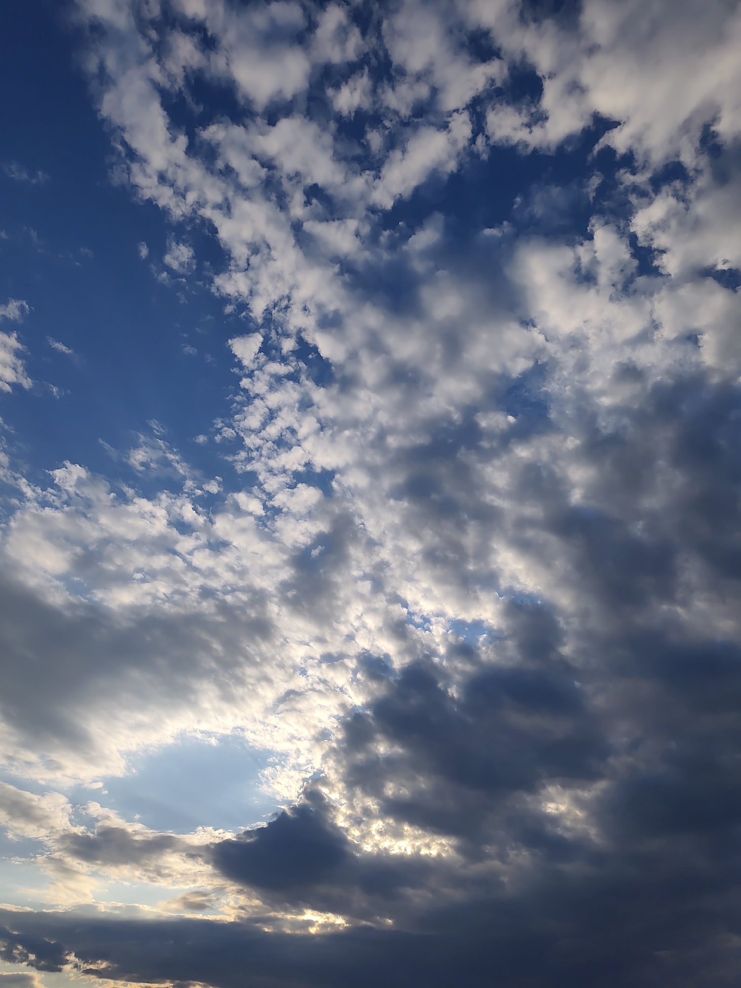 This is a photograph of a sky filled with clouds, viewed from the city of Codru, Moldova. The sky is predominantly blue with scattered white and dark gray clouds. Some of the clouds are illuminated by sunlight, creating bright patches and highlighting the textures of the cloud formations. The image appears to be taken during the daytime, possibly late afternoon or early evening, given the lighting. There are no people, buildings, or other man-made objects visible in the frame. The focus is entirely on the natural elements of the sky.