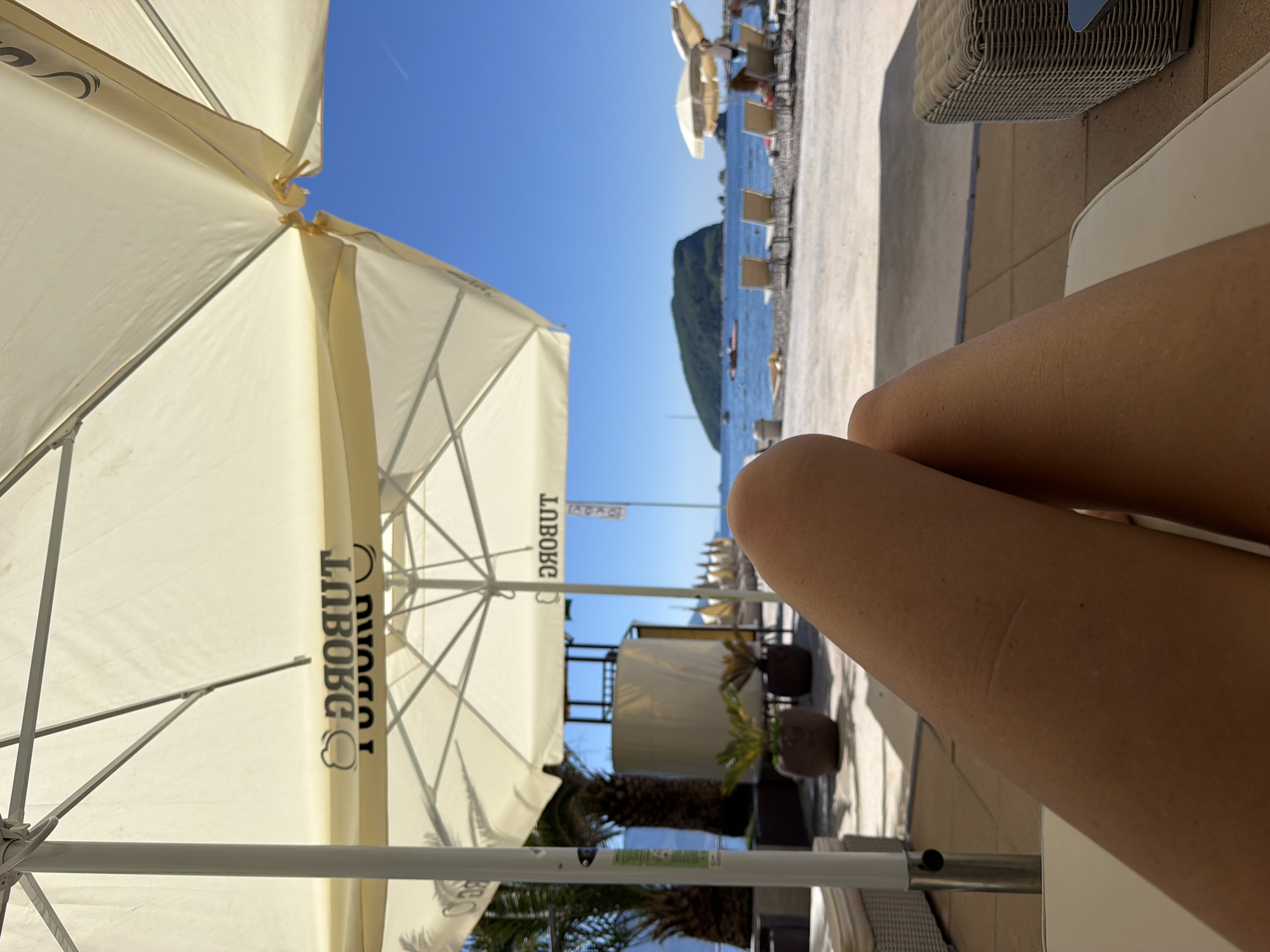 The image shows a beach scene with a view of the Adriatic Sea in Budva, Montenegro. Two fair-skinned legs are in the foreground, suggesting a person is relaxing on a beach chair. A beach umbrella with a 'LUBOG' sign obscures part of the scene. Further down the beach are more umbrellas and beach chairs. An island is visible in the distance, framed by a clear blue sky.